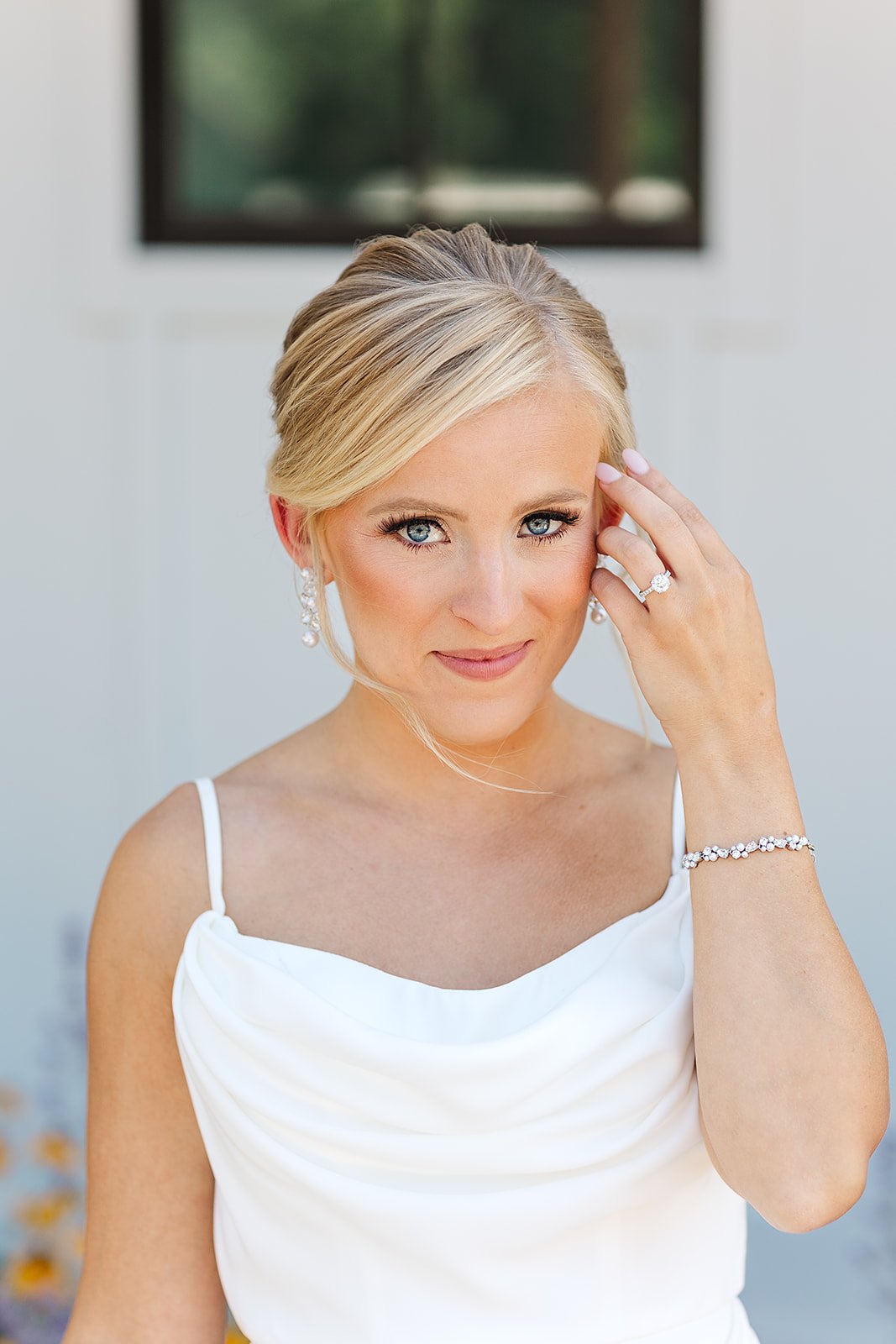A woman with blonde hair and blue eyes wearing a white dress, jewelry, and a wedding ring, standing outdoors with a blurred background and a window behind her.