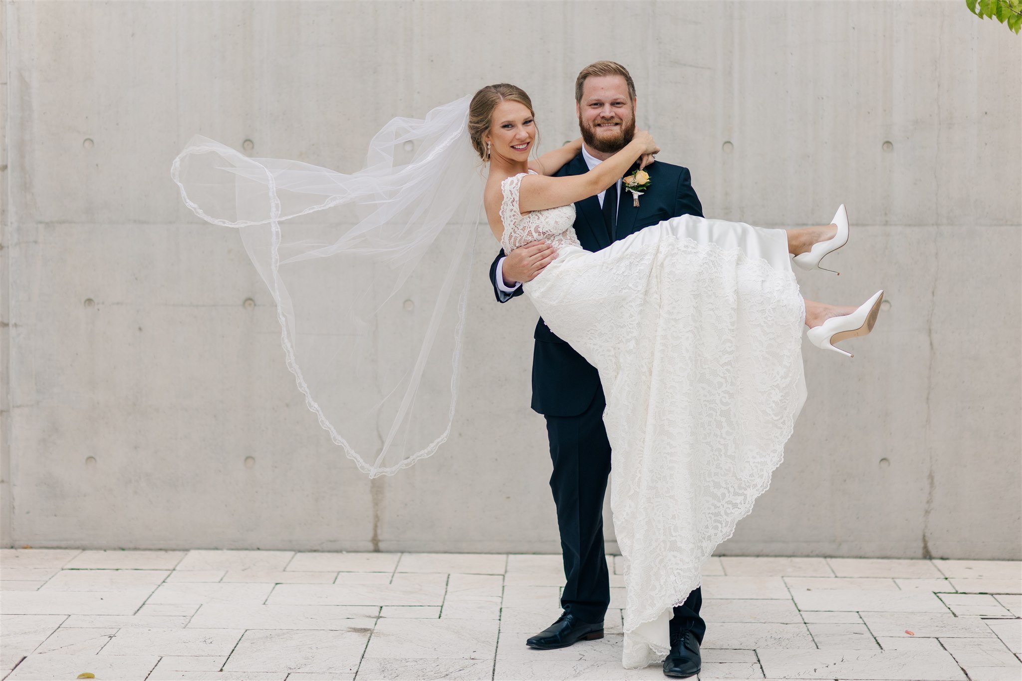 A bride and groom are standing in front of a concrete wall, with the groom holding the bride in his arms. The bride is wearing a white lace wedding dress and white high heels, and the groom is dressed in a dark suit with a boutonniere. The bride's lo