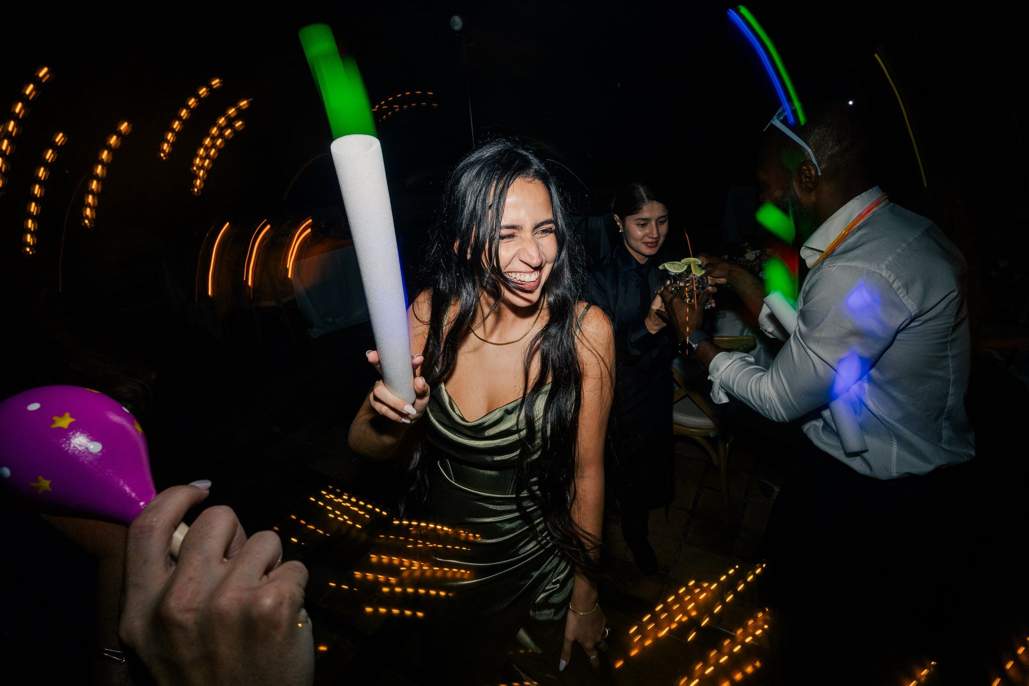 A woman with long dark hair smiling and holding a party noise maker at a lively celebration with colorful lights and other guests in the background.