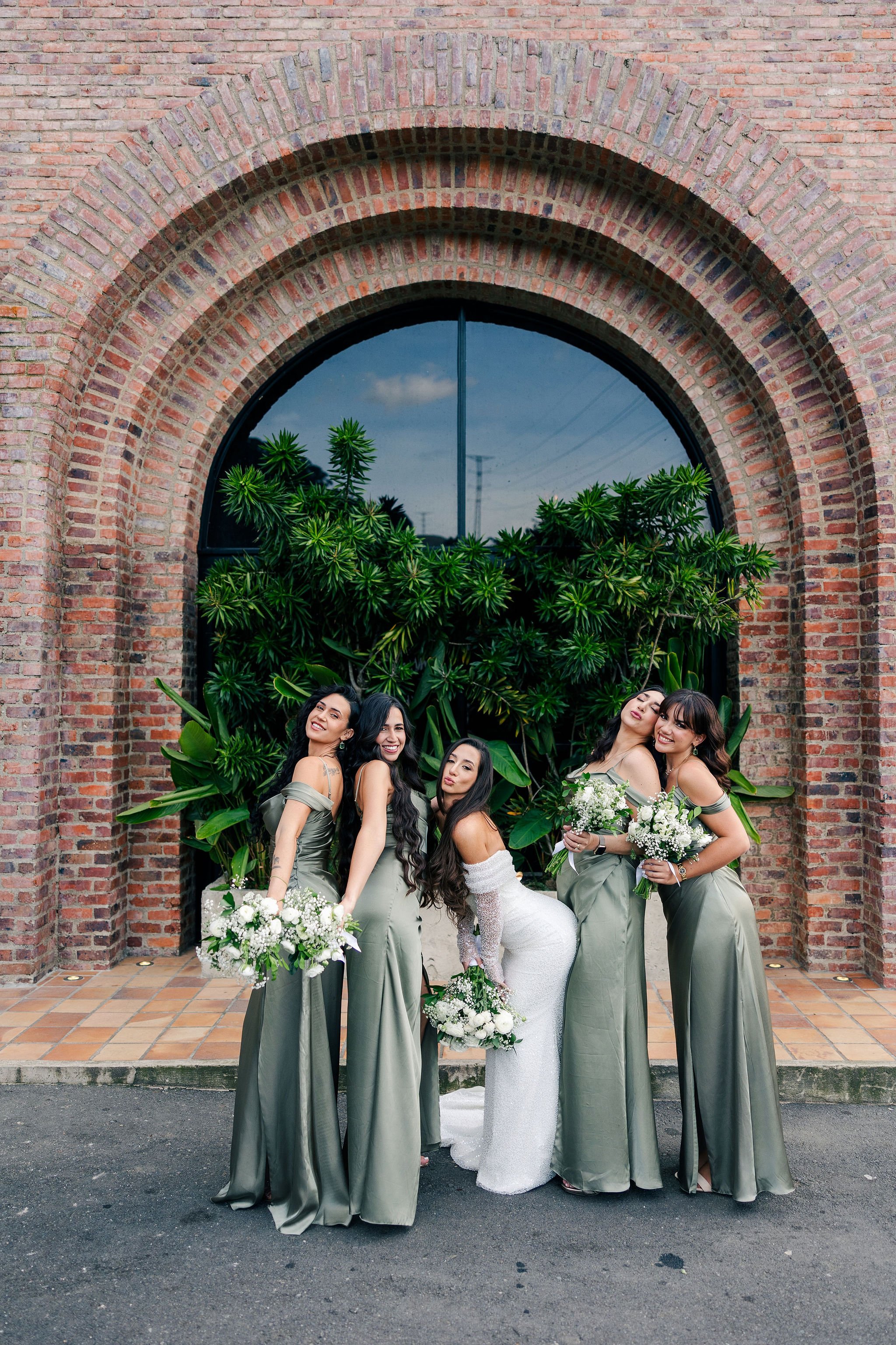 Bride in white wedding gown with four bridesmaids in matching olive green dresses holding bouquets of white flowers, standing in front of a brick archway and lush green plants.