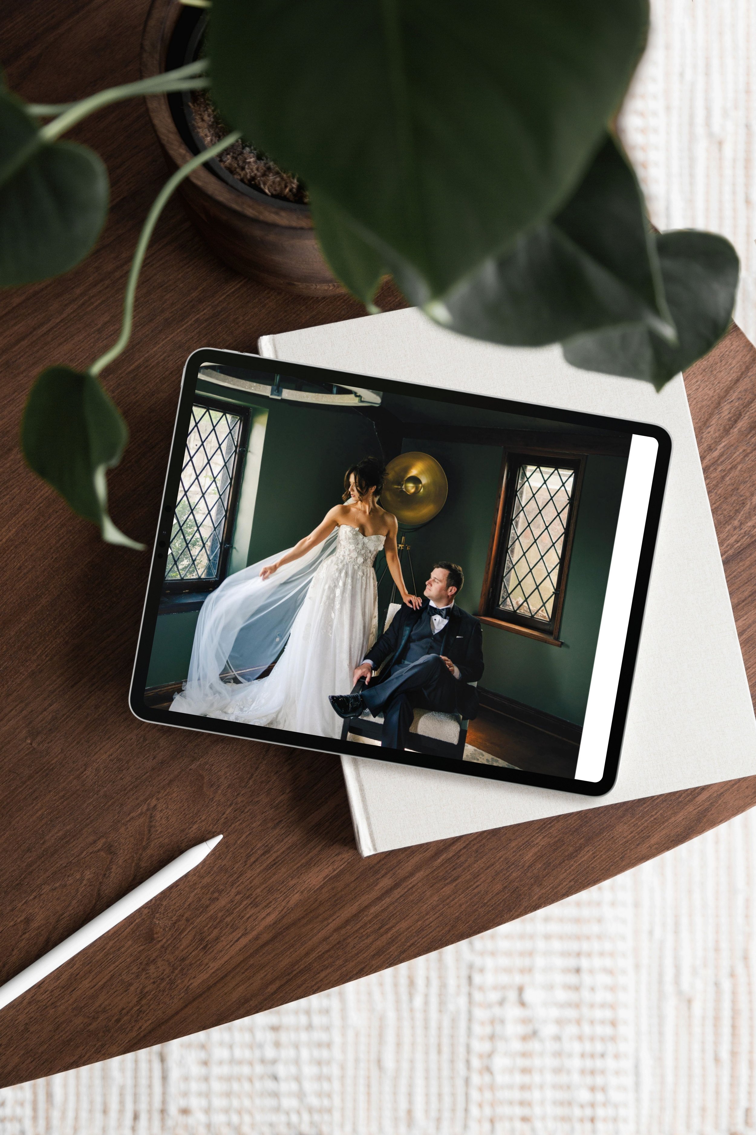 A tablet displaying a wedding photo of a bride in a white dress and a groom in a tuxedo inside a room with green walls, two windows, and a gold wall sculpture, placed on a white book on a wooden table, with a green plant and a white stylus nearby.