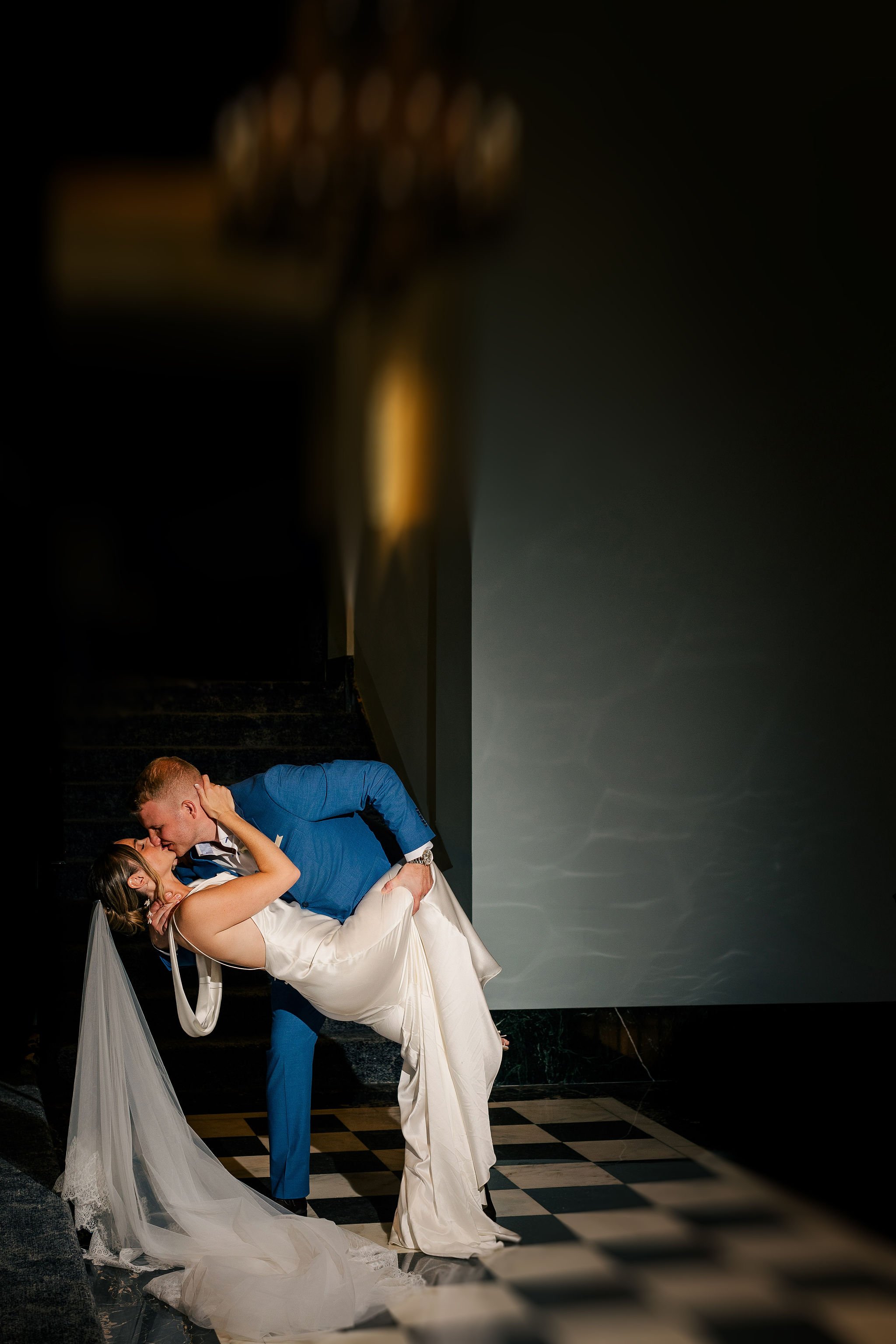 A newlywed couple sharing a romantic kiss in a dramatic pose on a checkered floor, with the groom leaning and dipping the bride, in front of a dark wall with a chandelier overhead.