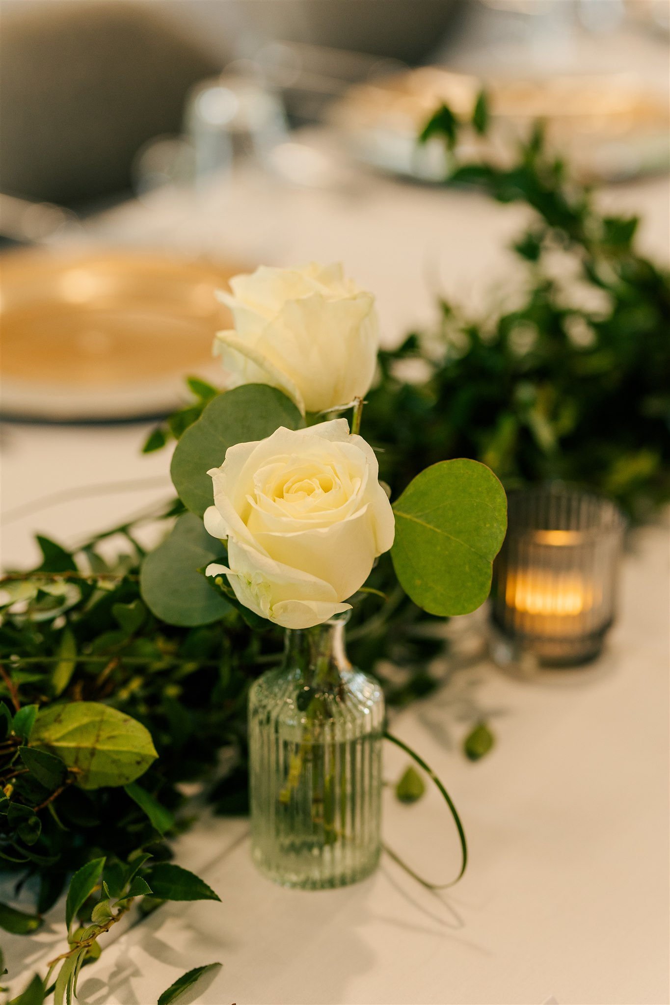 White roses in a small ribbed glass vase on a table decorated with greenery and a candle in a glass holder.