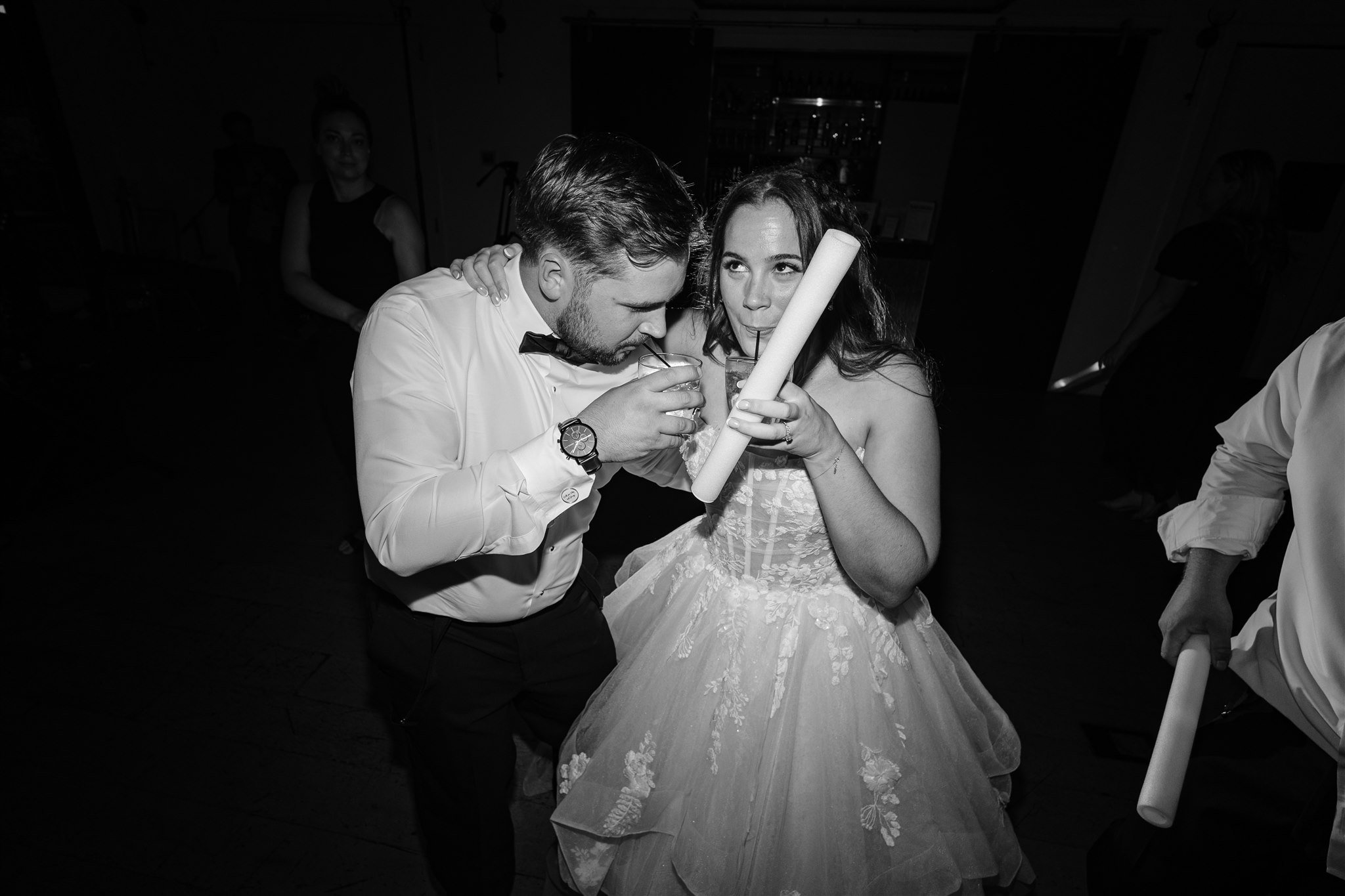 A bride and groom dance at a wedding reception, sharing drinks with straws while holding a party noisemaker.