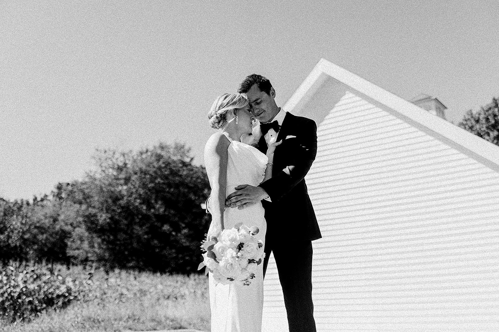 Black and white photo of a bride and groom embracing outdoors during their wedding, with a house in the background.