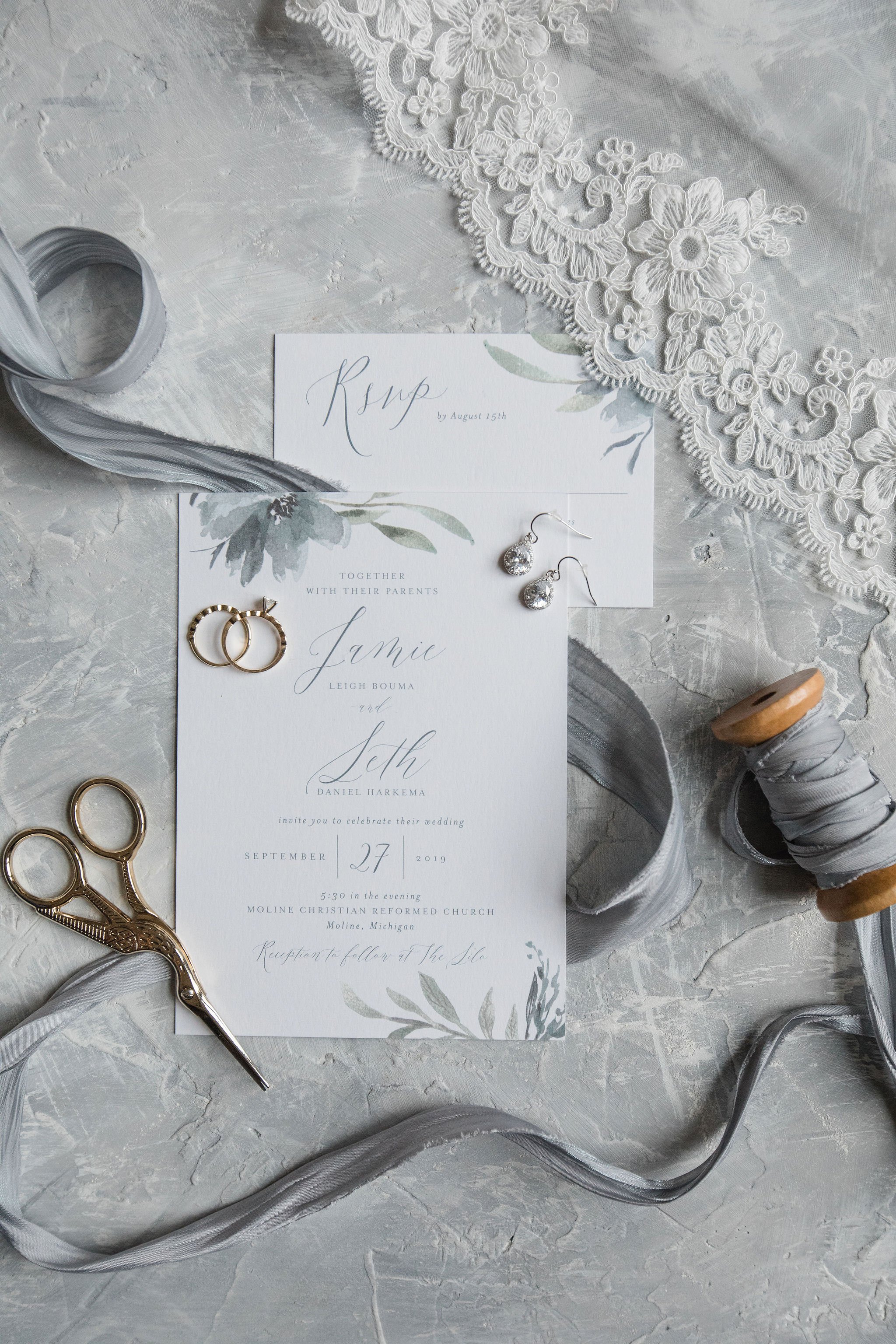Wedding invitation along with jewelry, scissors, ribbon, and lace fabric on a textured surface.