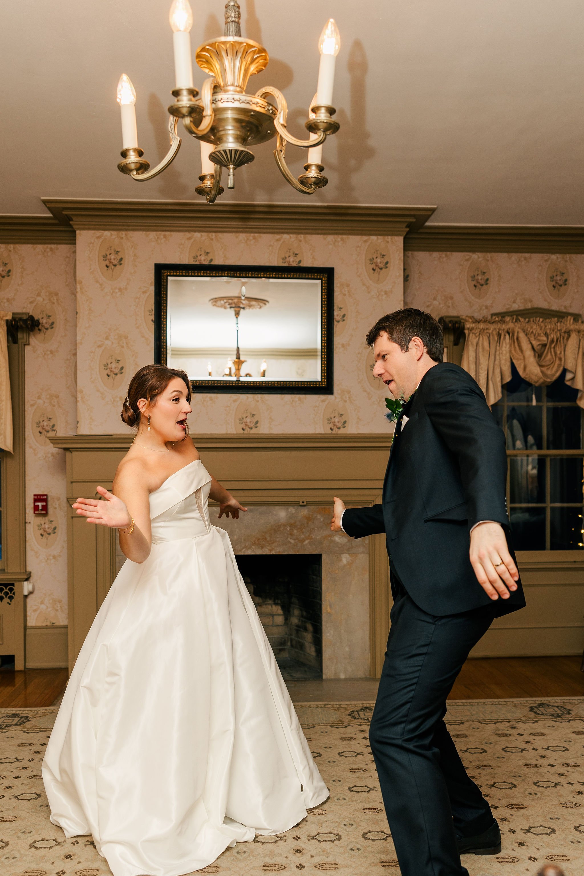 Bride and groom dancing together at their wedding reception in a decorated room with a chandelier overhead and a mirror reflecting the ceiling light.