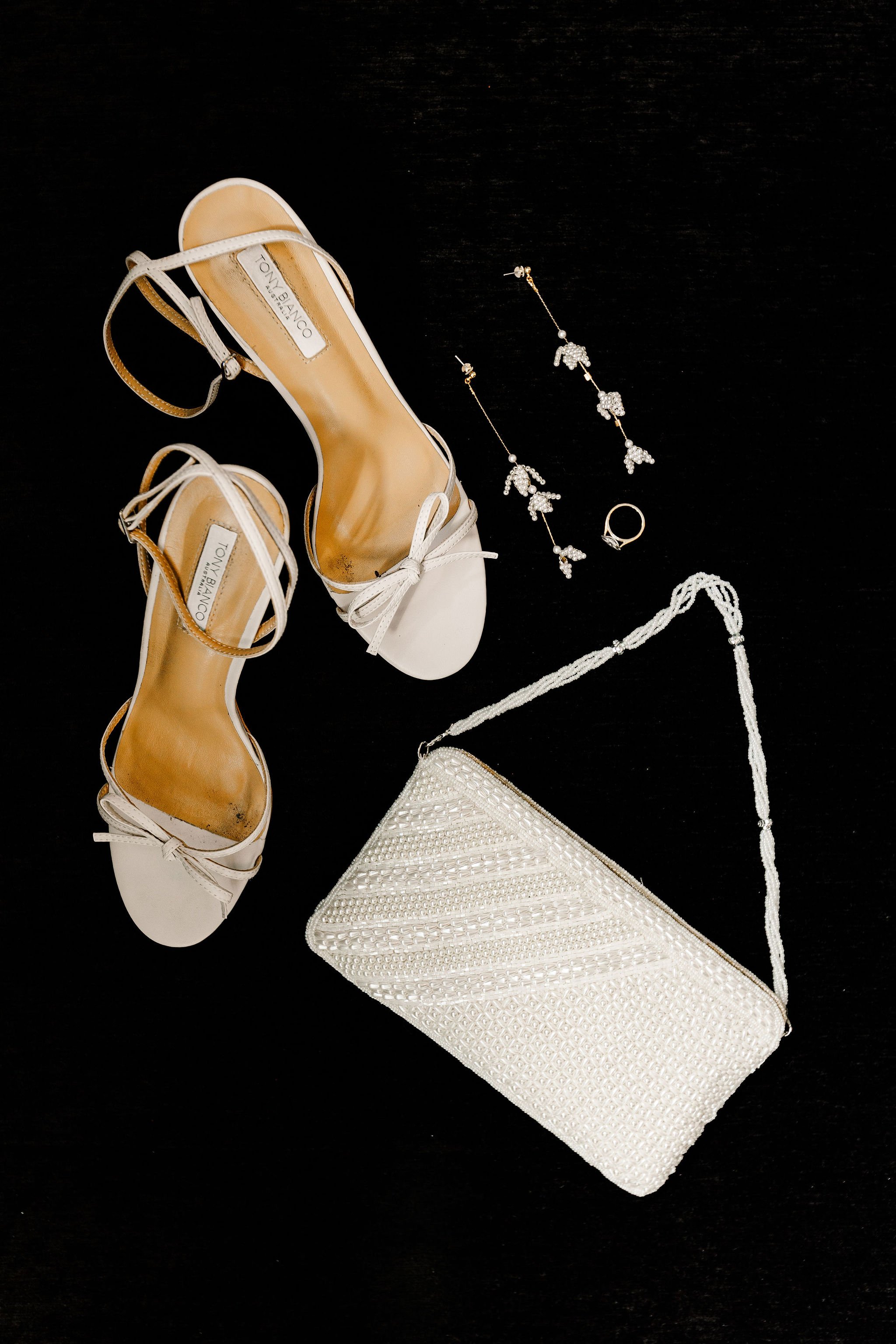 White high-heeled sandals, pearl clutch purse, matching pearl jewelry including earrings and a ring, all arranged on a black surface.