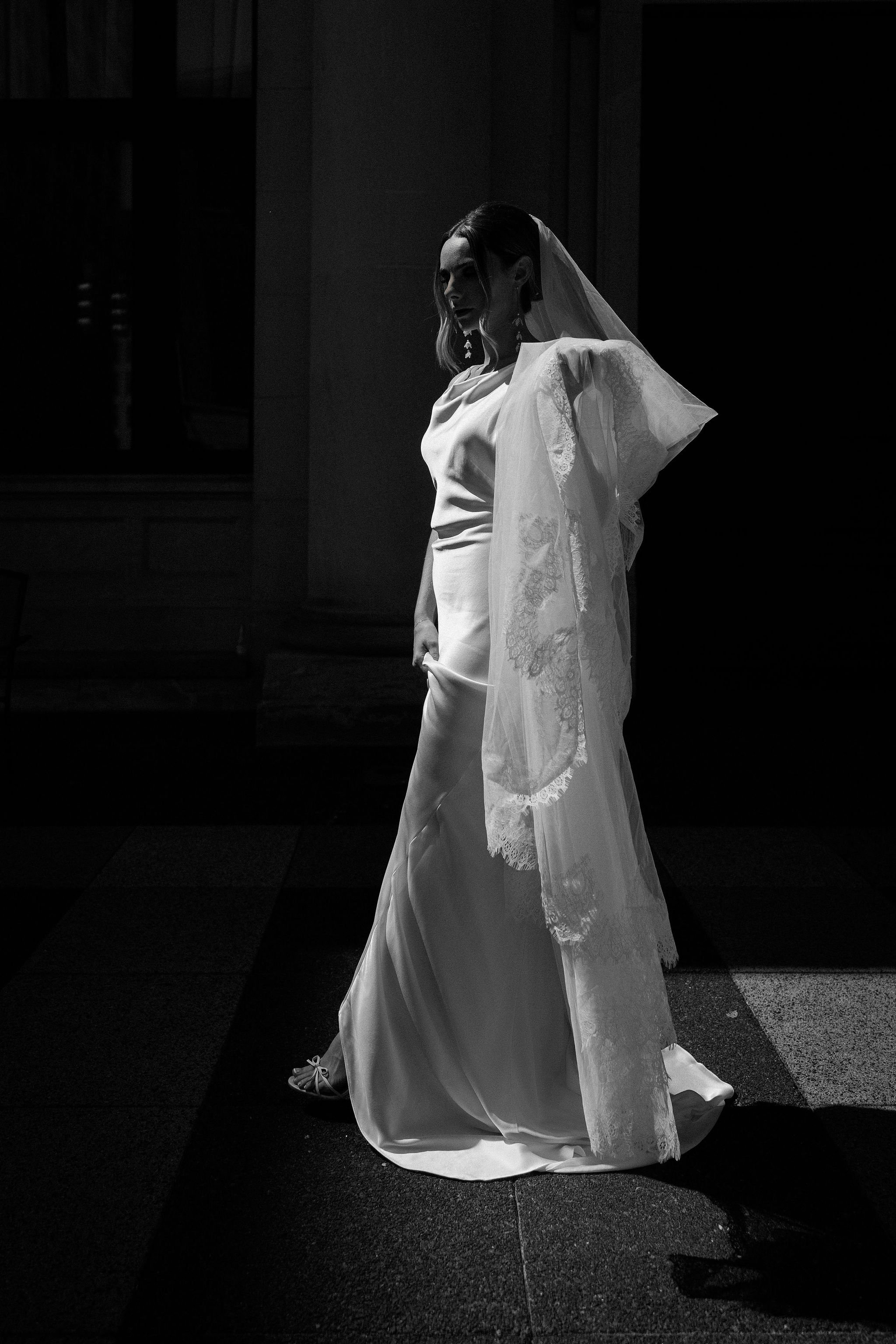 A woman in a wedding dress and veil stands in a low-lit setting, with a focused light illuminating her from the front.