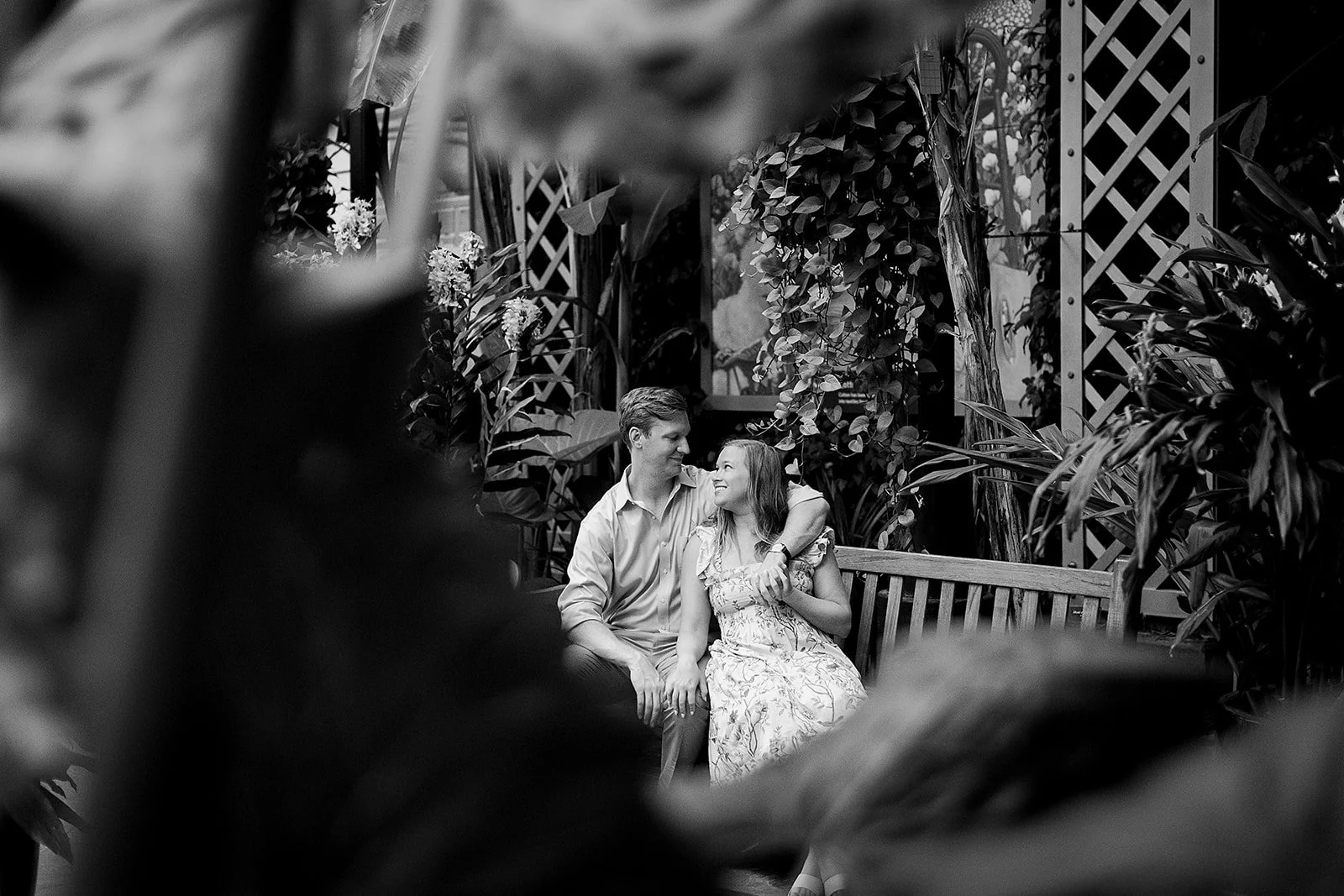 A black and white photo of a man and woman sitting on a park bench surrounded by plants, gazing at each other and smiling.
