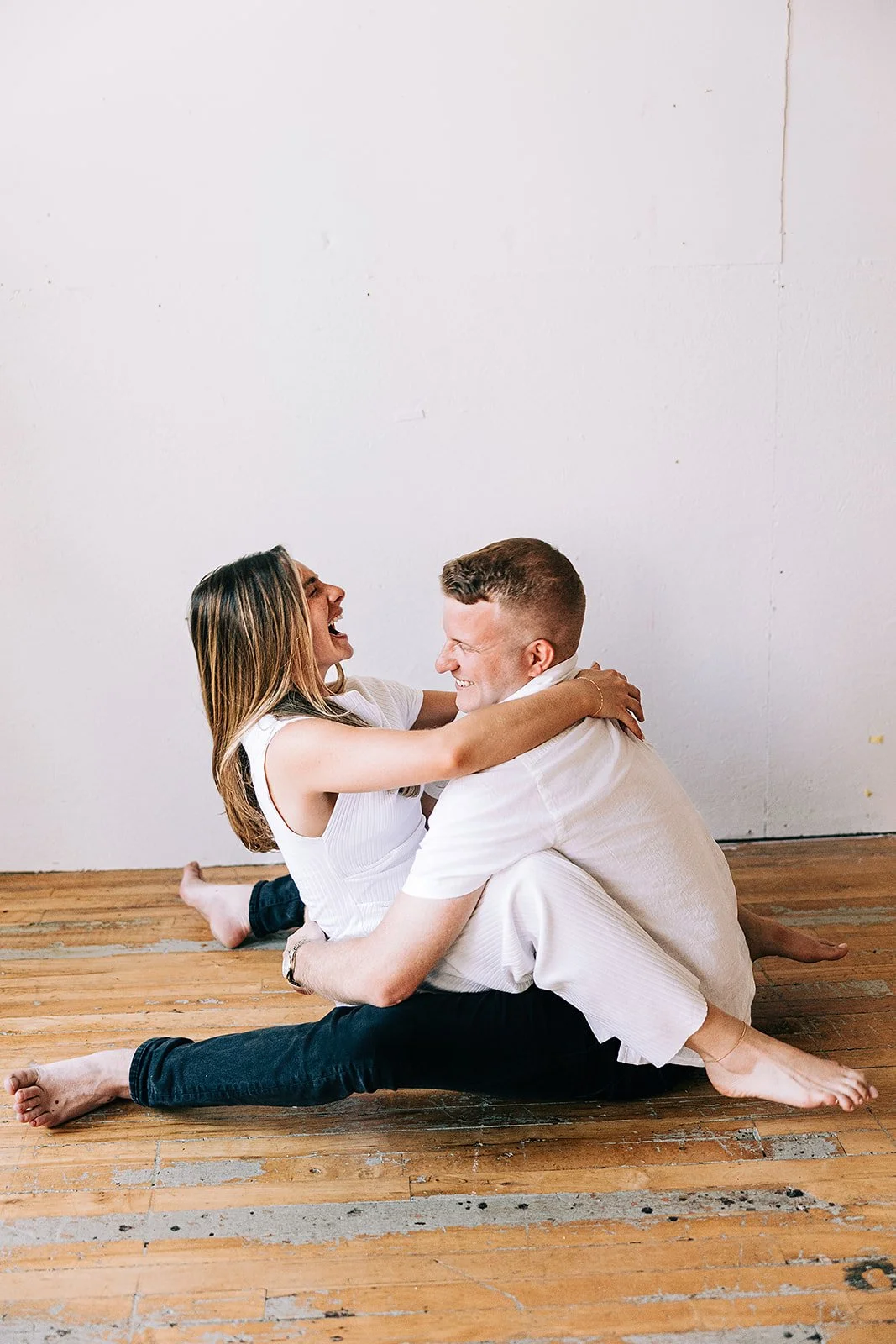 A couple sitting on a wooden floor, hugging and smiling at each other, against a plain white wall.