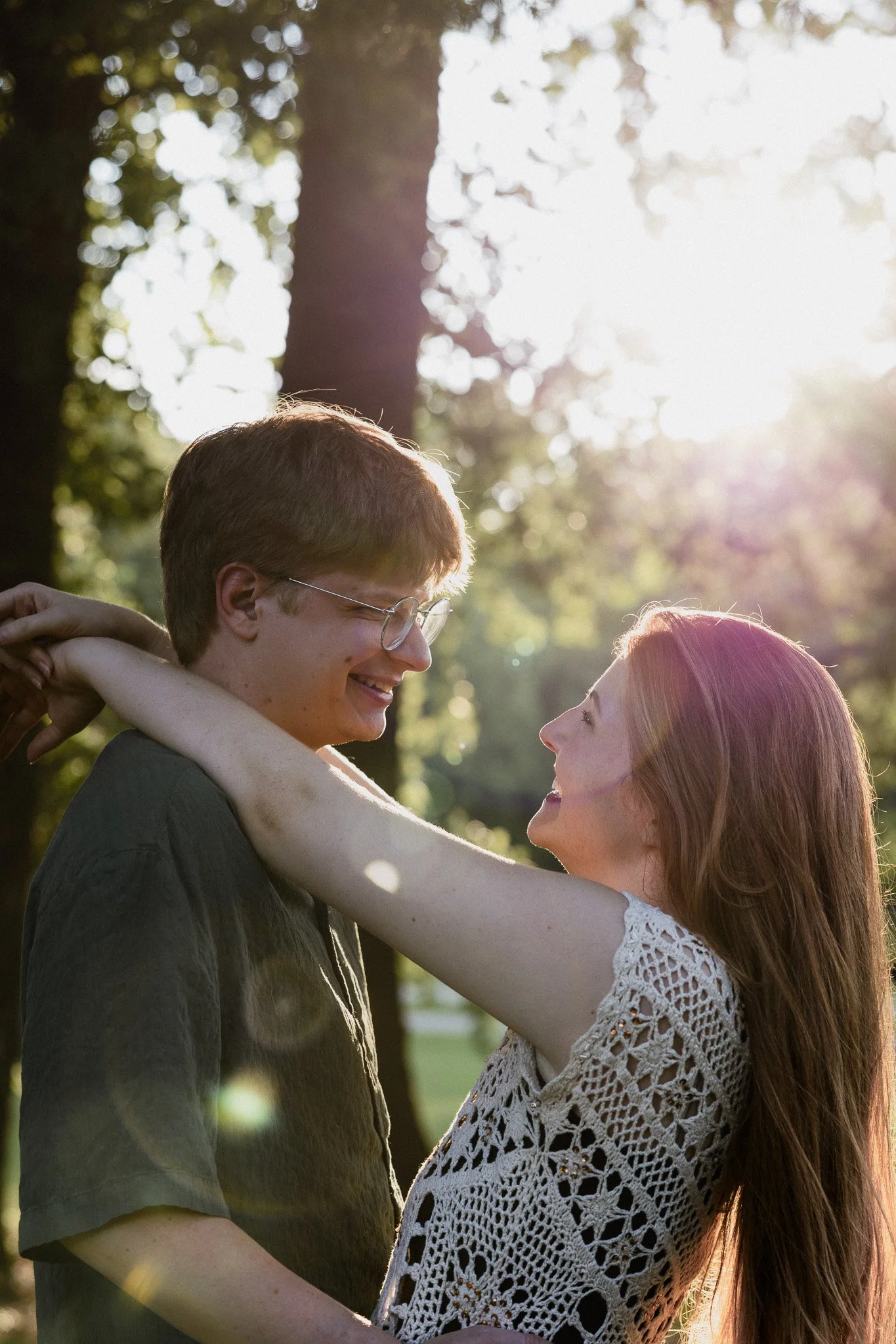Couple embracing and smiling at each other during an outdoor couples photography session, with warm sunlight filtering through the trees.