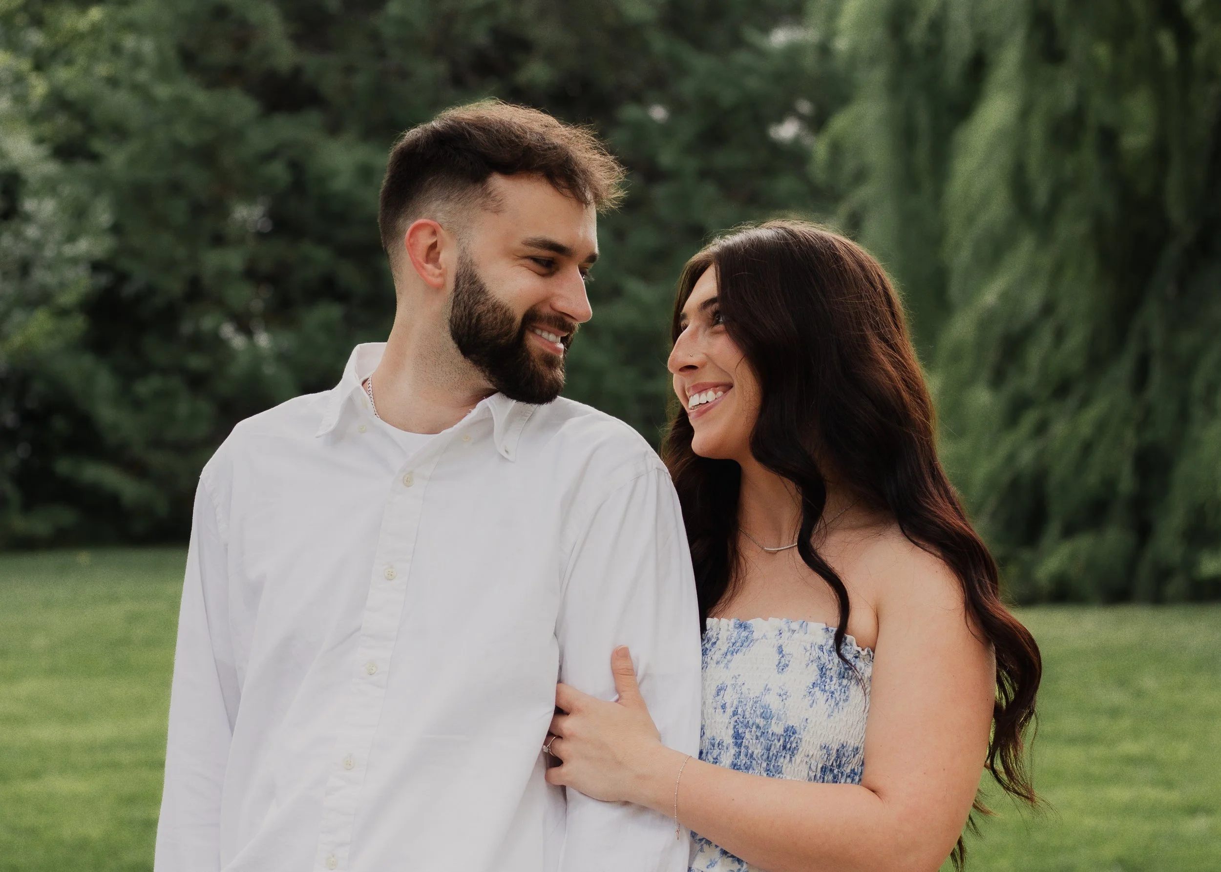 A smiling man and woman look at each other in a park with lush green trees and grass.
