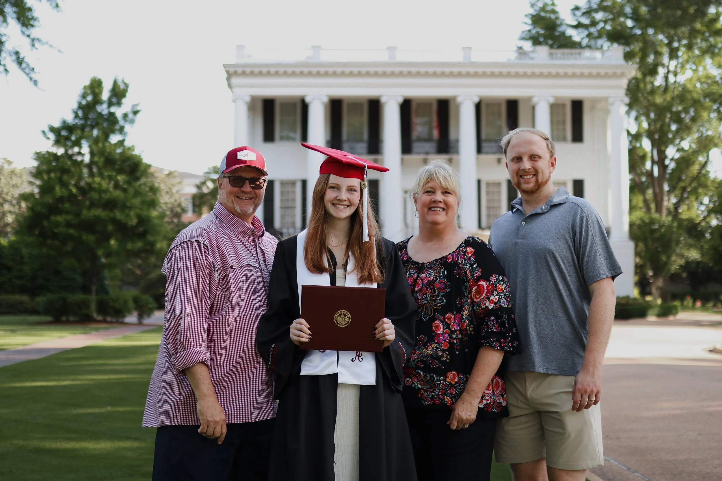 college graduate standing outside in cap and gown with her family holding degree