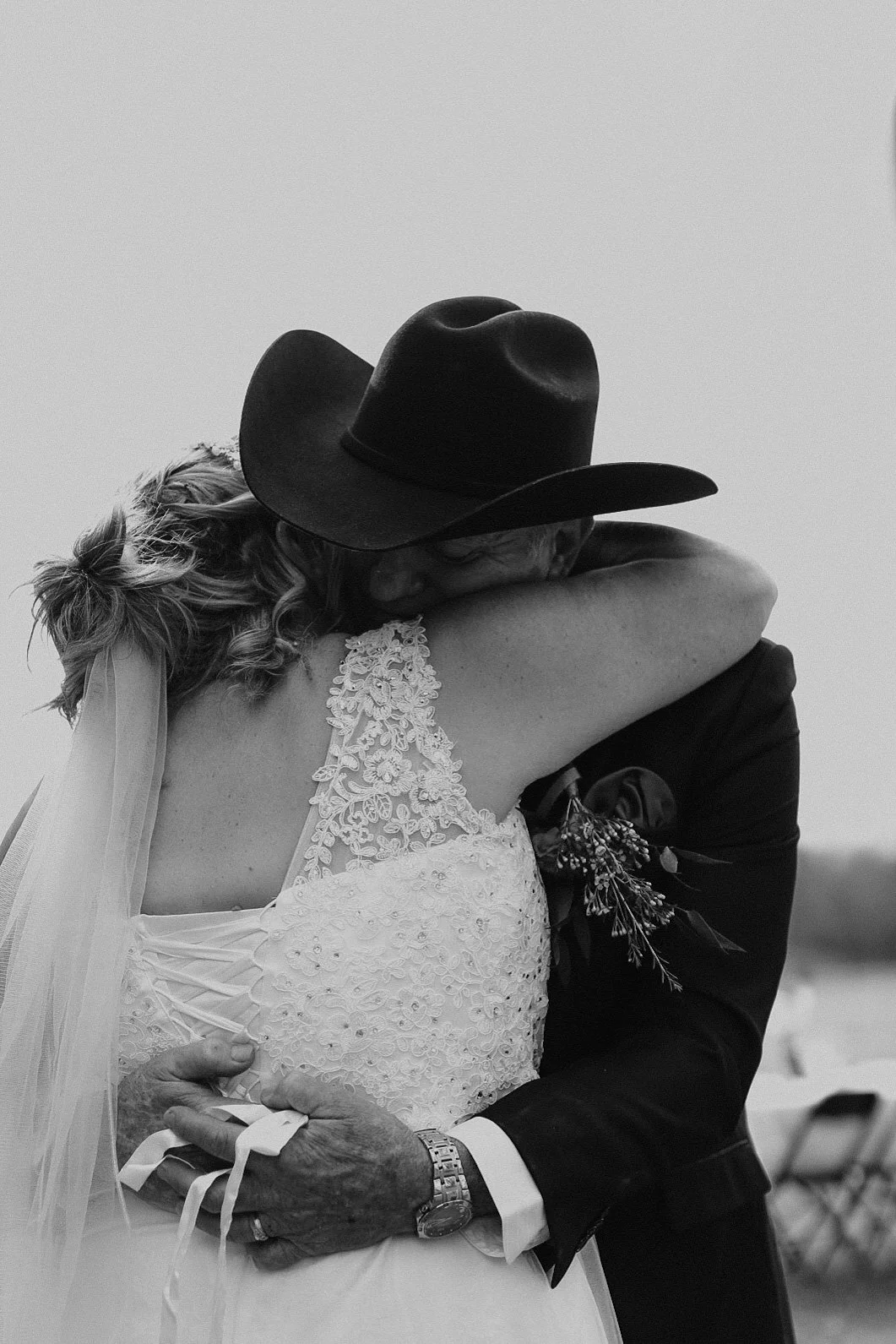 A bride and groom embrace during their wedding, with the bride wearing a lace wedding dress and veil, and the groom dressed in a dark suit and cowboy hat.