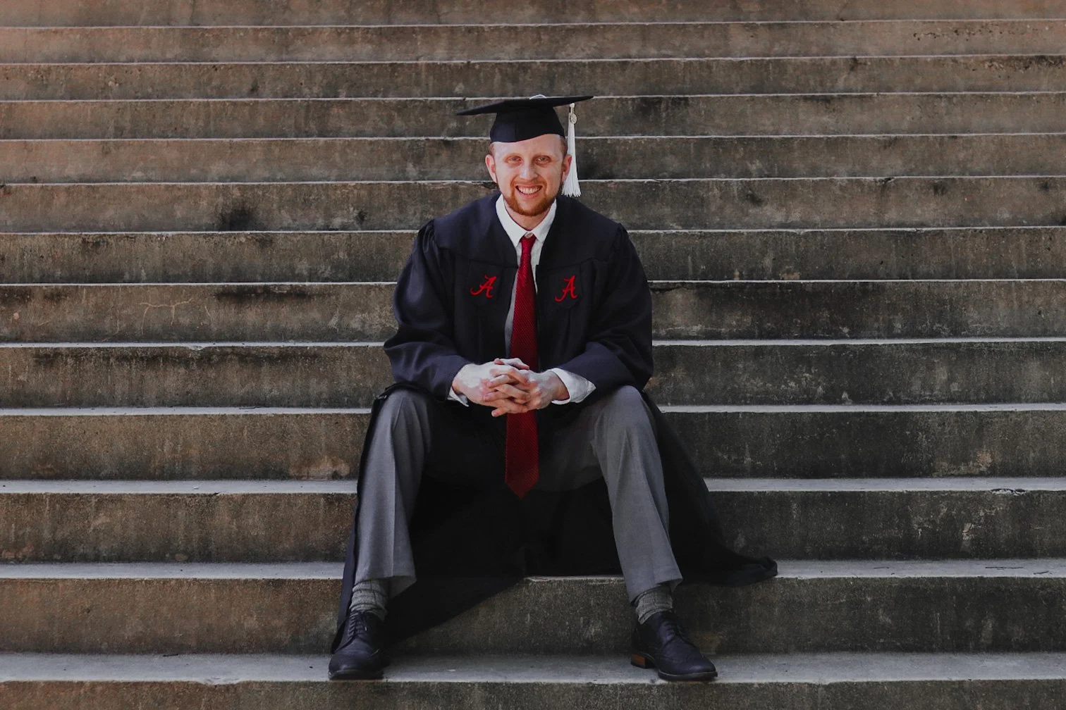 male college graduate sitting outside on steps in cap and gown