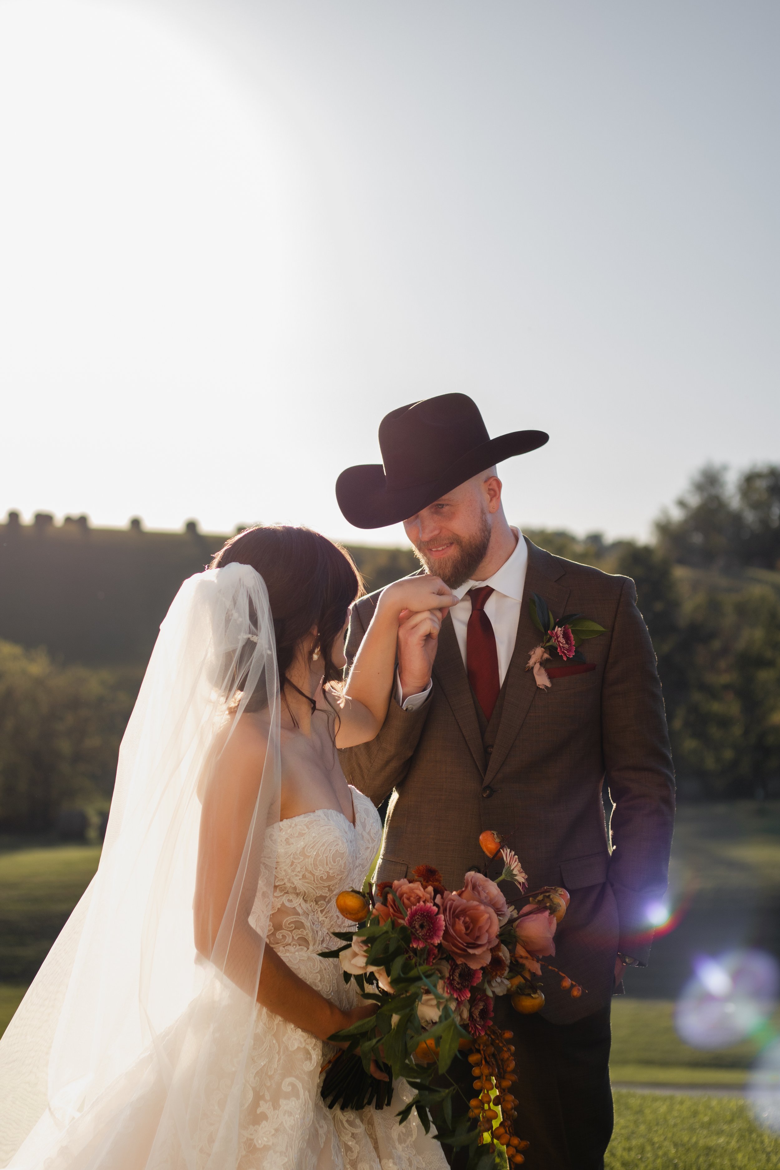 A bride and groom sharing a tender moment outdoors during their wedding, with the groom wearing a cowboy hat and suit, and the bride holding a bouquet of flowers.