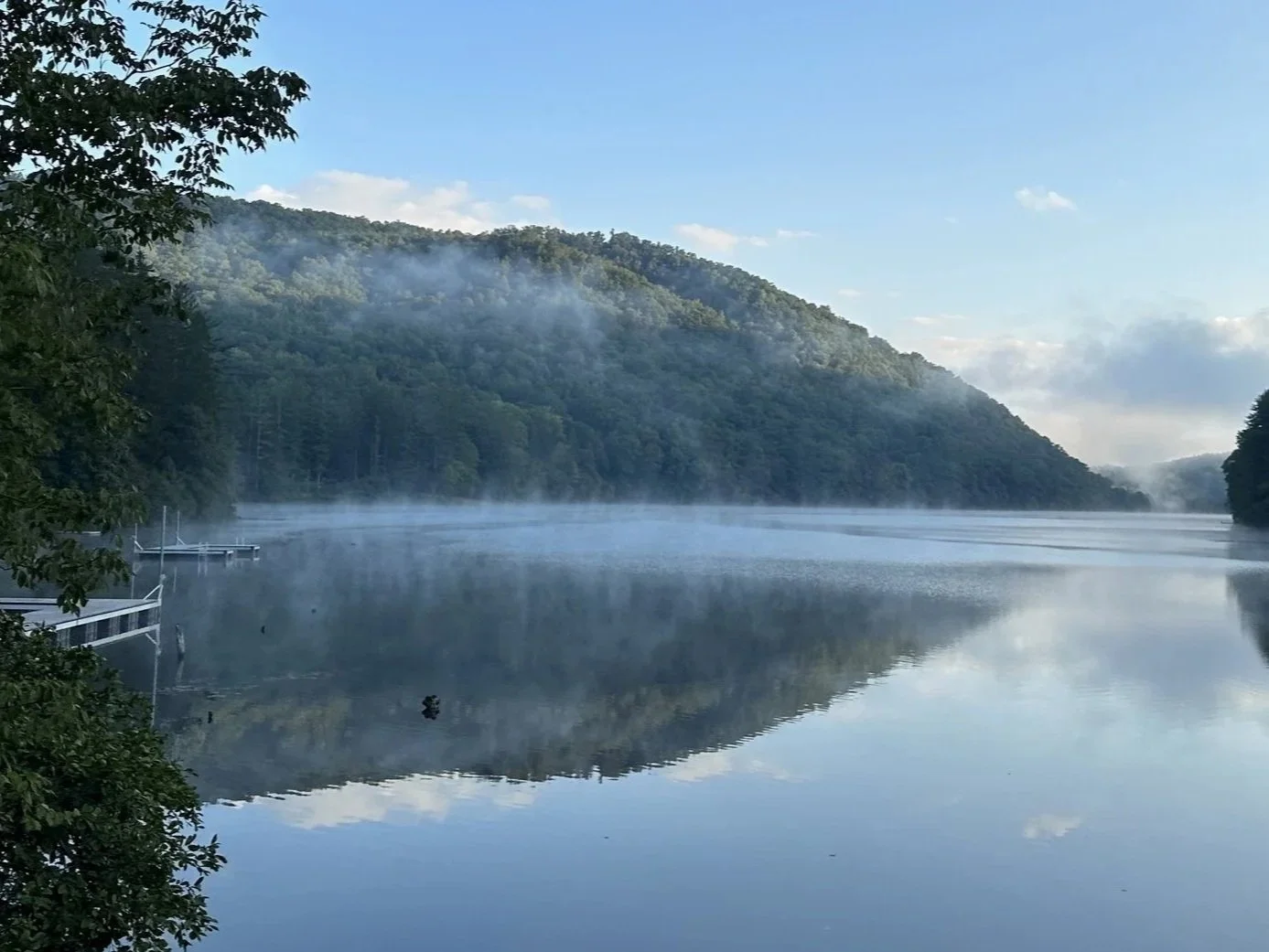 Calm river with a mist rising above the water, surrounded by forested hills under a clear blue sky.