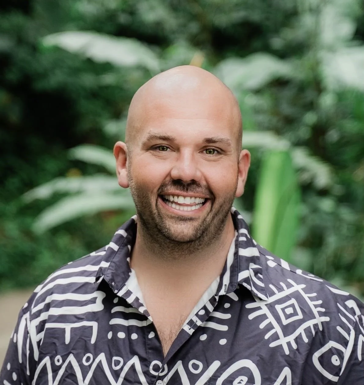 Photo of Jake Sasseville, a white man wearing a patterned collared shirt and smiling at the camera. He is bald and has light facial stubble.. There are plants in the background.