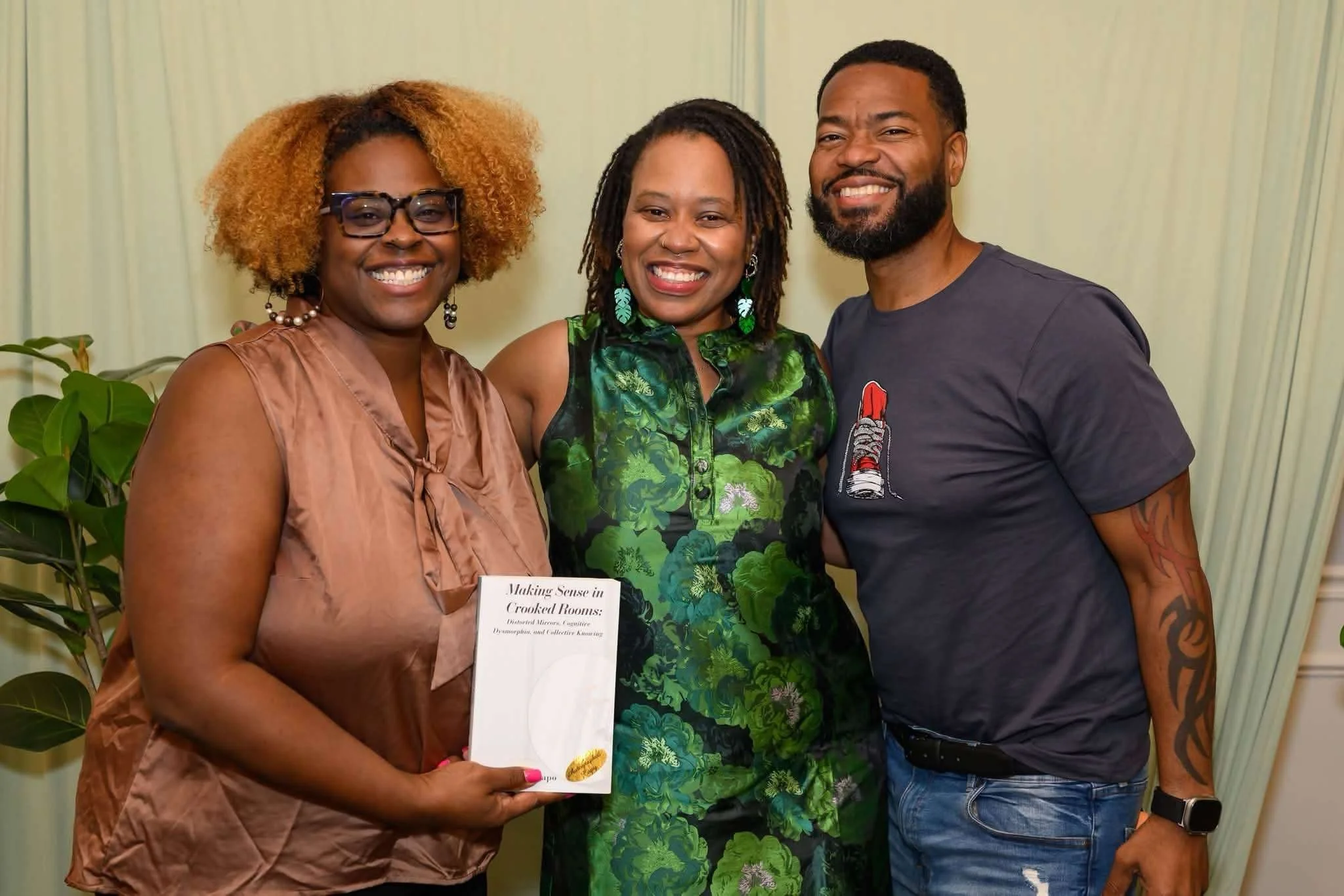 Jessica, a Black woman wearing a green dress, stands with two other people at her book launch event. They are holding a copy of her book, and all of them are smiling widely.
