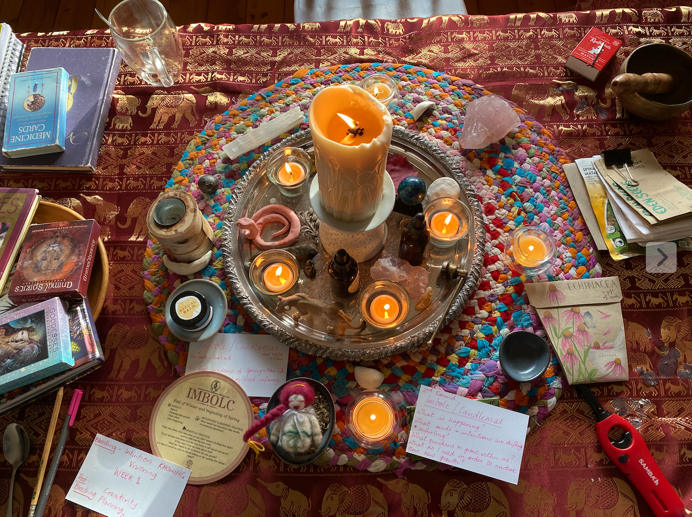 A colorful altar setup with a large wax candle, smaller candles, crystals, small bottles, and decorative items on a silver tray, surrounded by books, notes, and various objects on a patterned cloth.