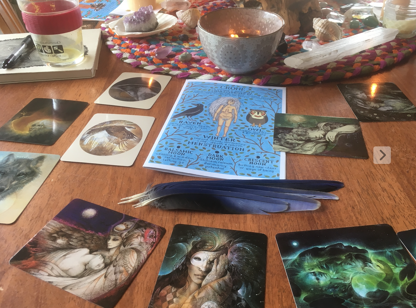 A table set up for a tarot or oracle card reading session. In the center, there is a blue pamphlet with mystical symbols and references to lunar phases. Spread out on the table are various tarot or oracle cards with artistic, fantasy-style illustrations, a small pile of blue and white feathers, a candle, a bowl, a jar of crystals or stones, and other small items. The background shows a colorful woven cloth with shells, crystals, and other decorative objects.