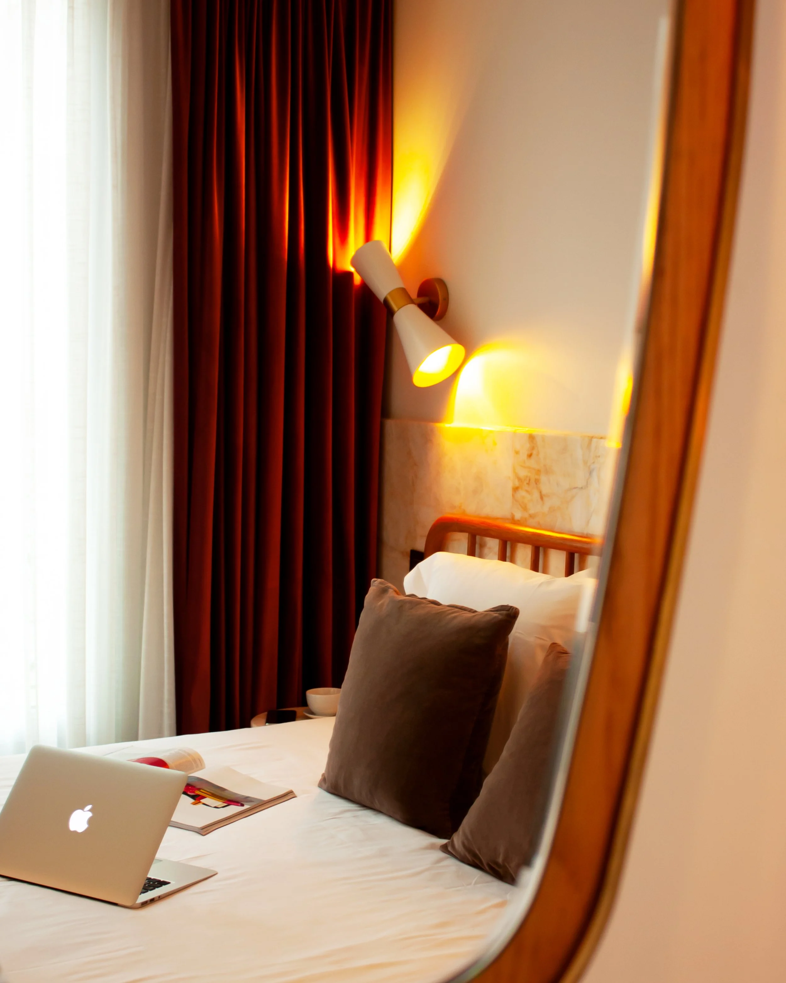 Cozy bedroom with a neatly made bed, laptop, magazine, pillows, modern wall lamp, and curtains.