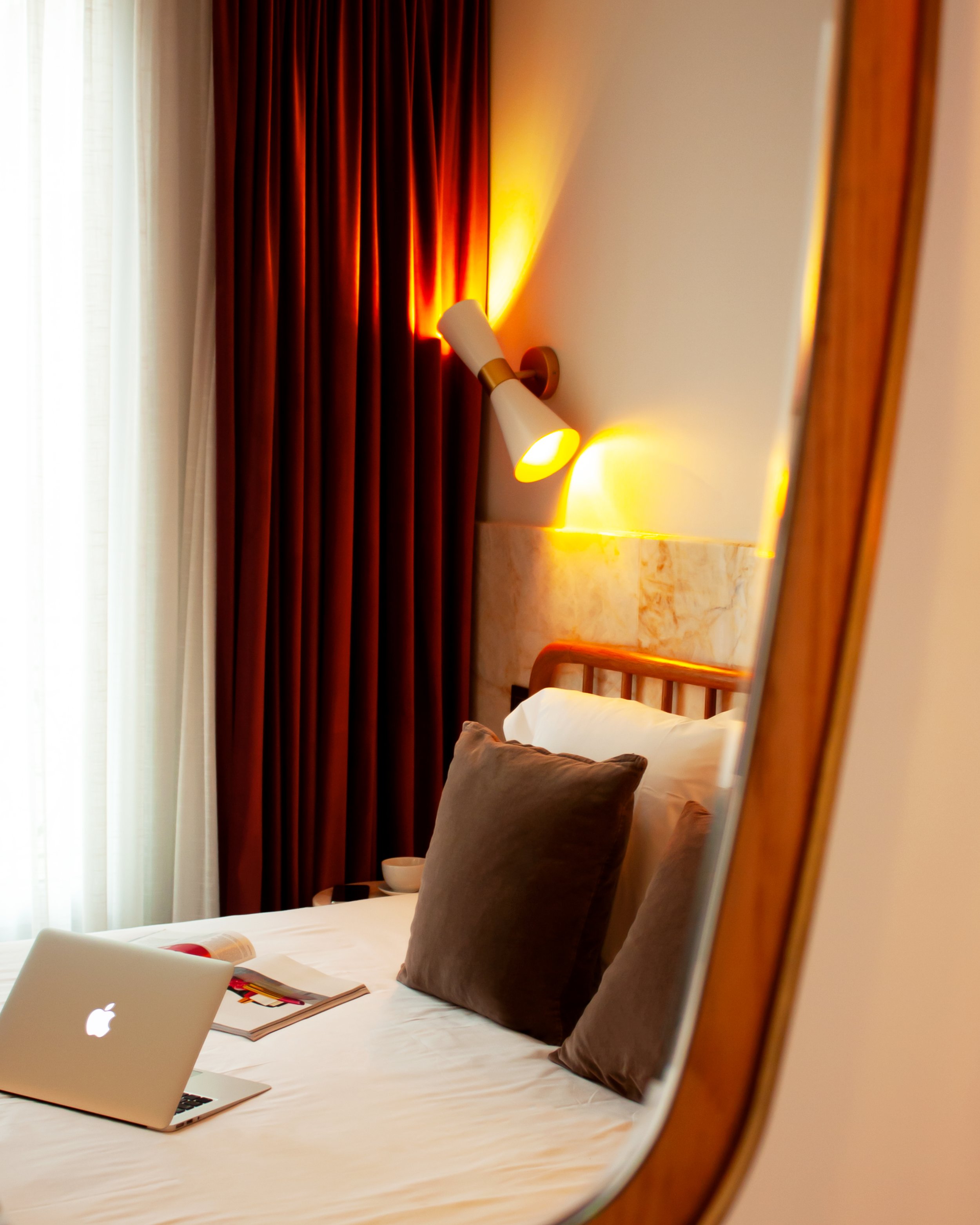 Cozy bedroom with a bed featuring white linens and brown pillows, a laptop, and an open magazine. There are maroon curtains, a wall-mounted lamp, and a mirror reflecting the scene.