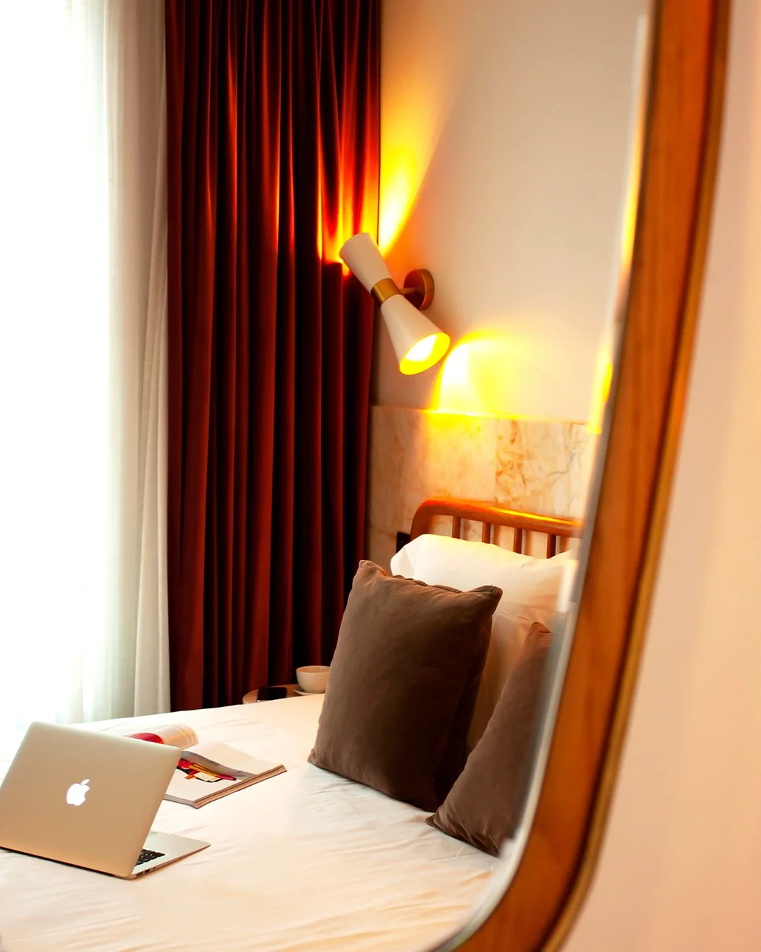 A cozy hotel room with a neatly made bed featuring two brown pillows. A laptop and magazine are placed on the white bedding. Warm lighting from a wall-mounted lamp and red curtains create a comfortable atmosphere.