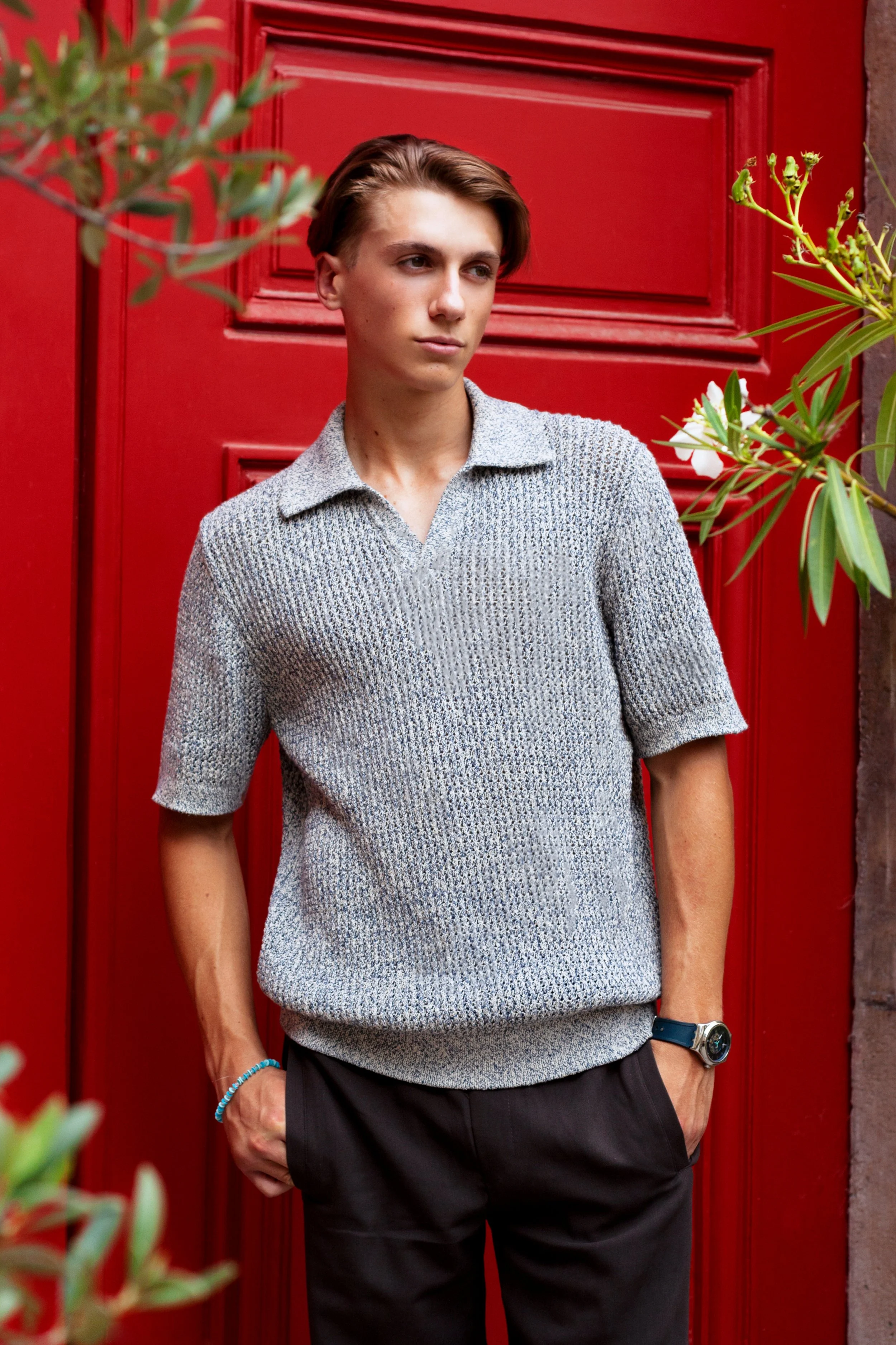 Person wearing a gray knit shirt standing in front of a red door with green plants nearby.