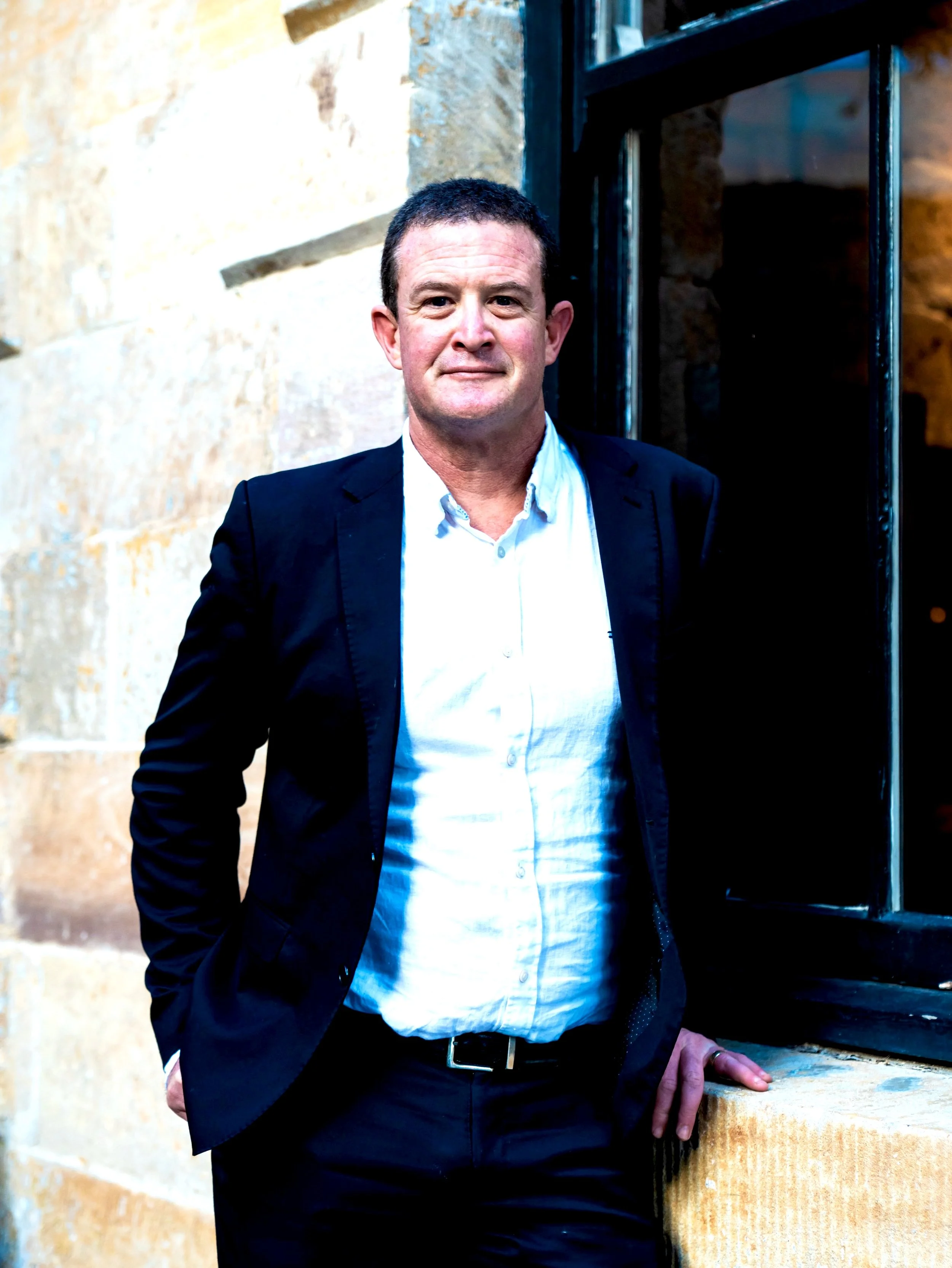 A man in a black suit and white shirt standing against a brick wall and window.