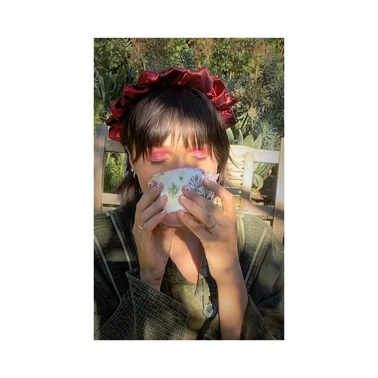 A person with short dark hair, wearing a red shiny headband, enjoys a drink from a beautiful hand-crafted porcelain chicken tea cup with floral-patters in an outdoor garden setting.