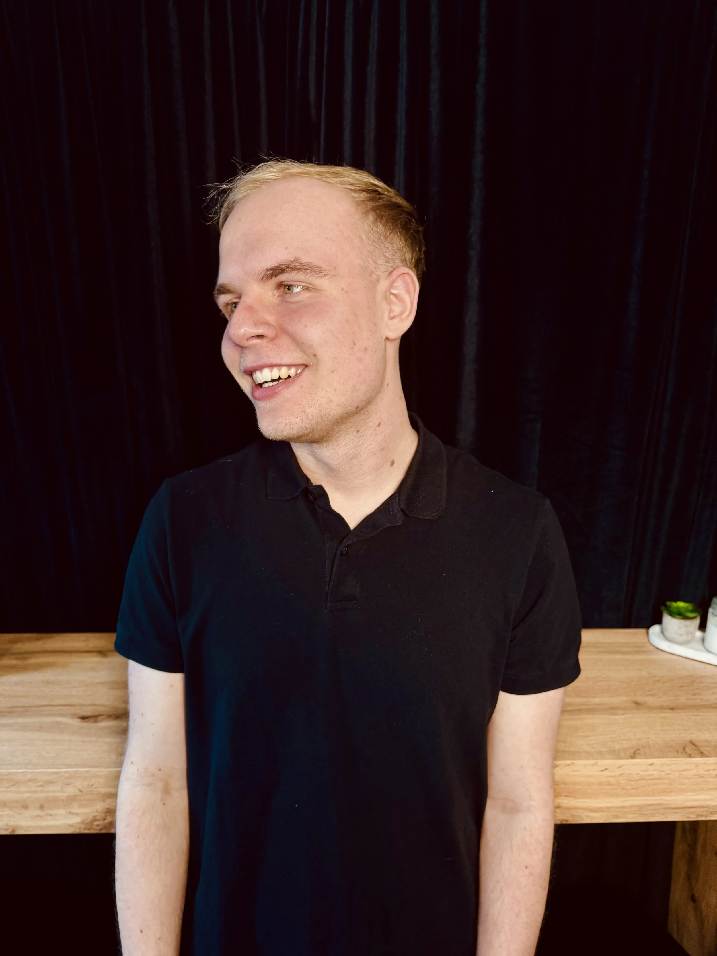 A young man with short blonde hair, wearing a black polo shirt, smiling and looking to the side, standing in front of a black curtain with a wooden table behind him.