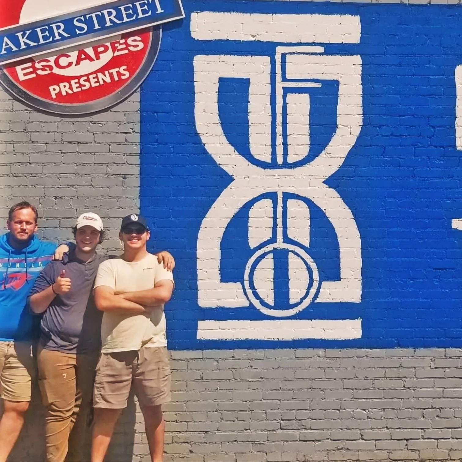 Three young men standing in front of a painted brick wall featuring a blue and white basketball-themed mural and a sign reading 'Baker Street ESCAPES presents' for an escape room in downtown Oklahoma City.