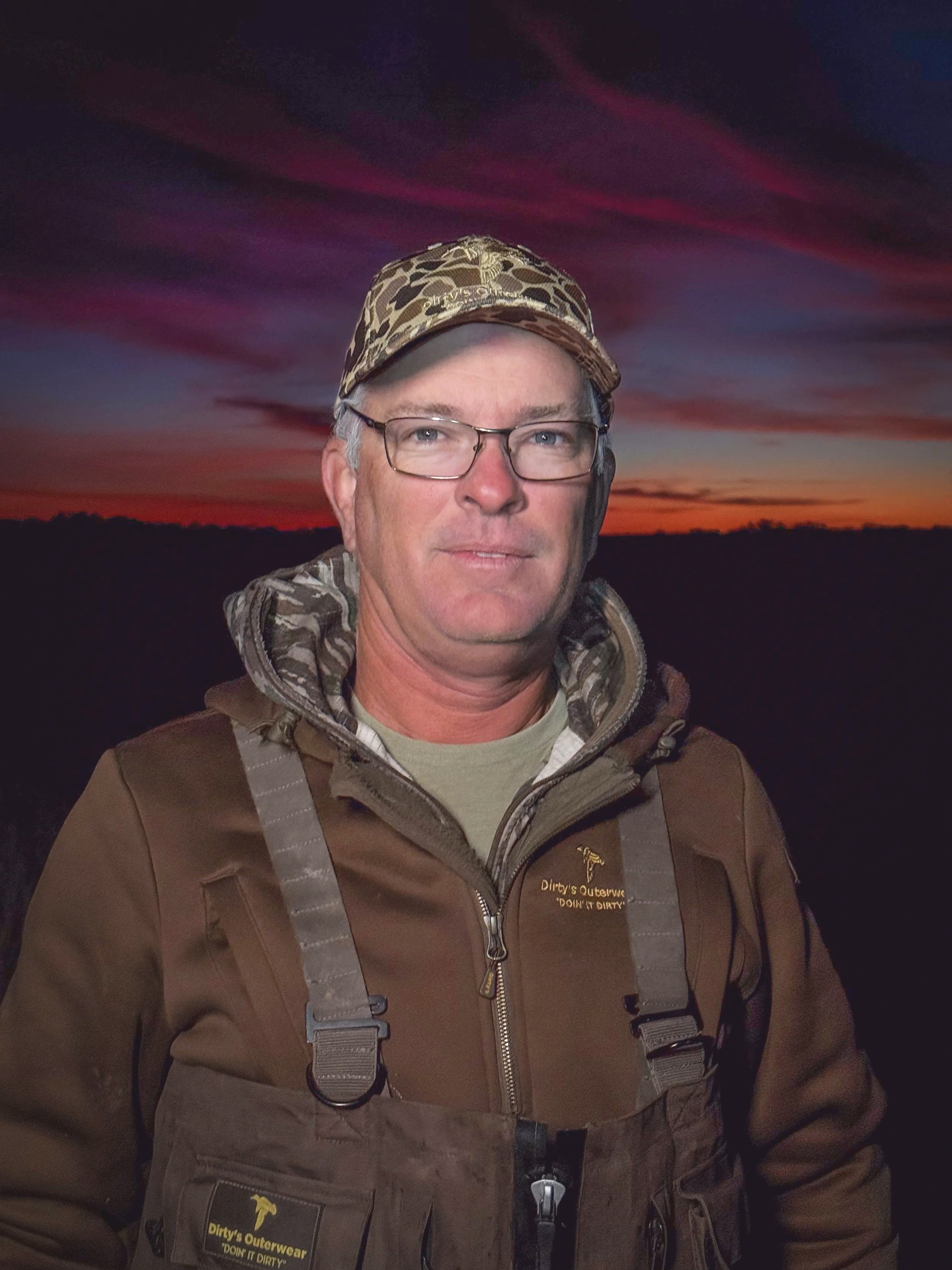 A man wearing glasses, camouflage hat, and brown outdoor gear stands outdoors at sunset with colorful sky in the background.