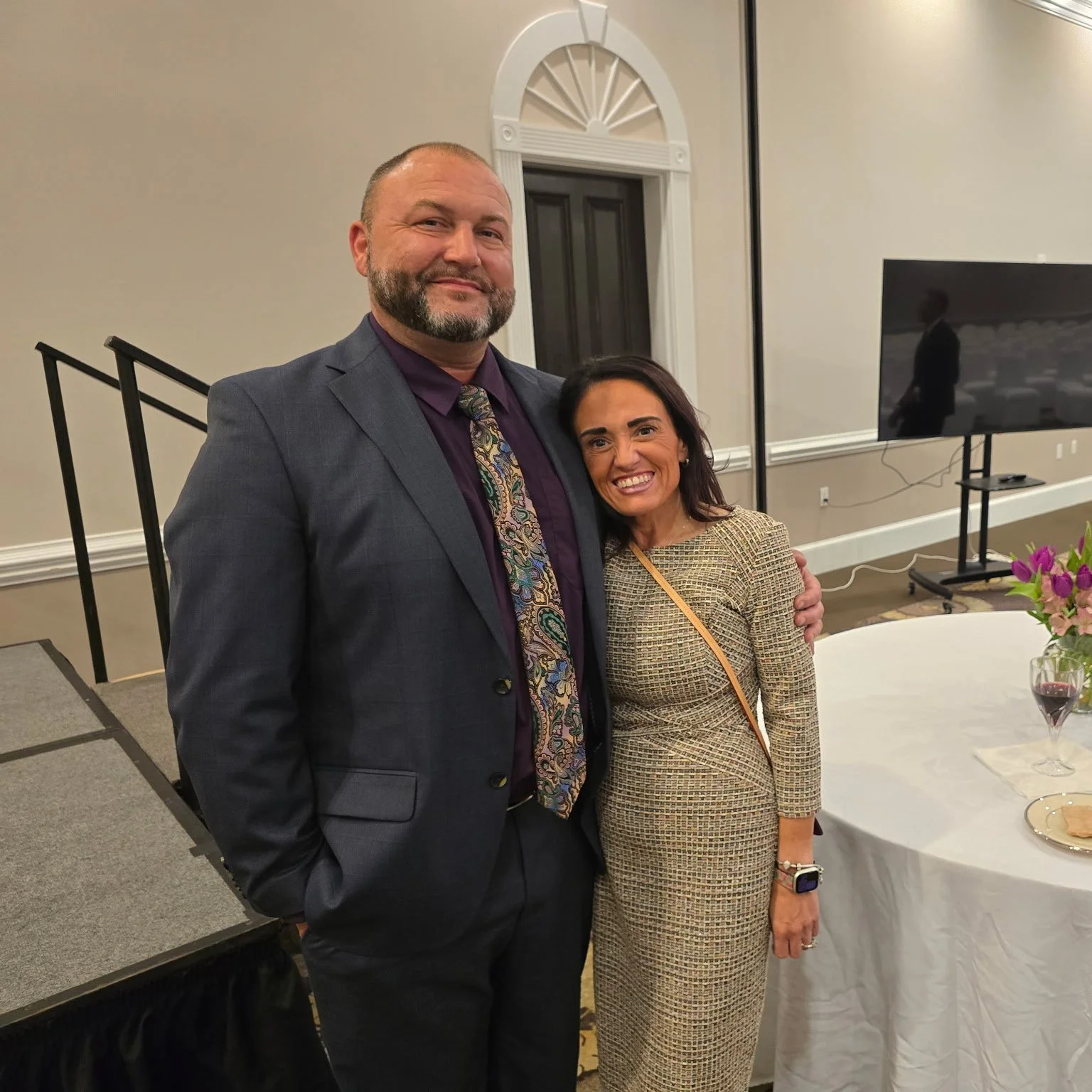 A man and woman smiling and posing together at an indoor event, with a table with flowers and a large TV screen in the background.