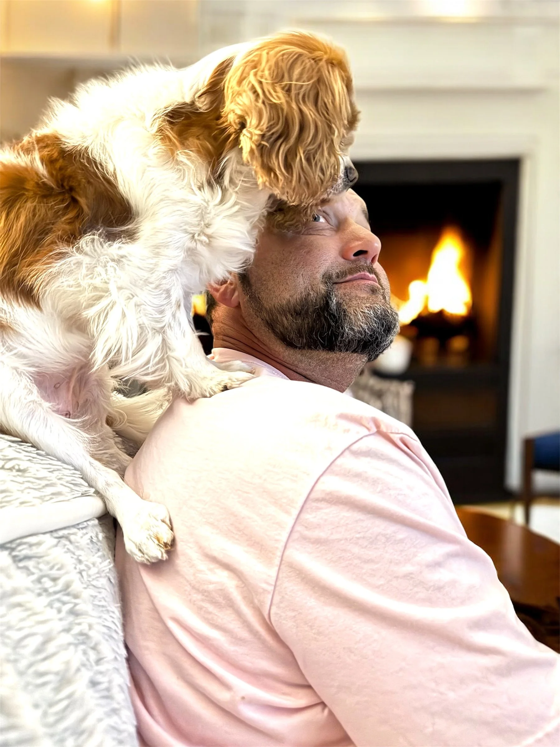 A man with a beard and a dog with long, floppy ears sitting on his shoulders in front of a fireplace with a fire.
