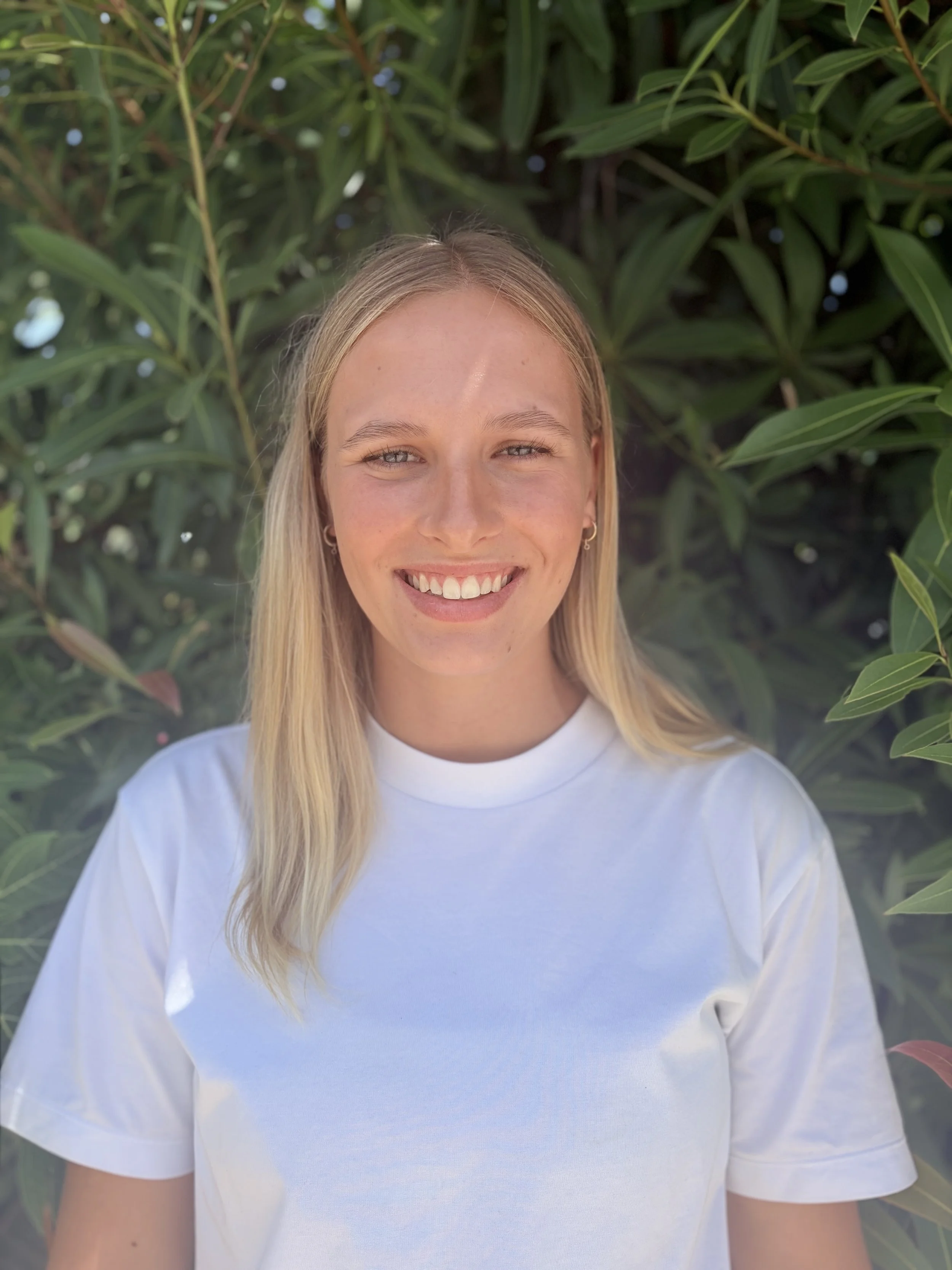 A female physiotherapist wearing a white T-shirt and smiling in front of some bushes