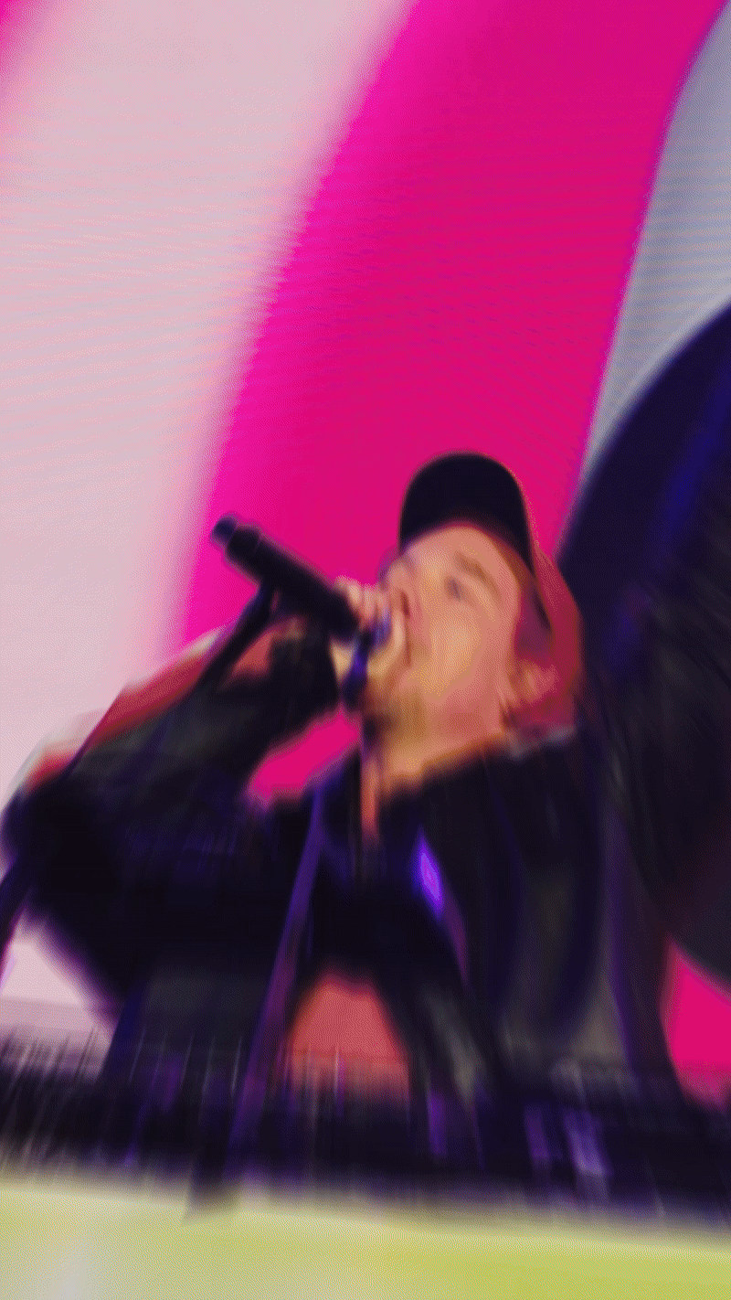 A person wearing a black jacket and a black cap holding a microphone, performing on stage with colorful lighting, and a blurred background.