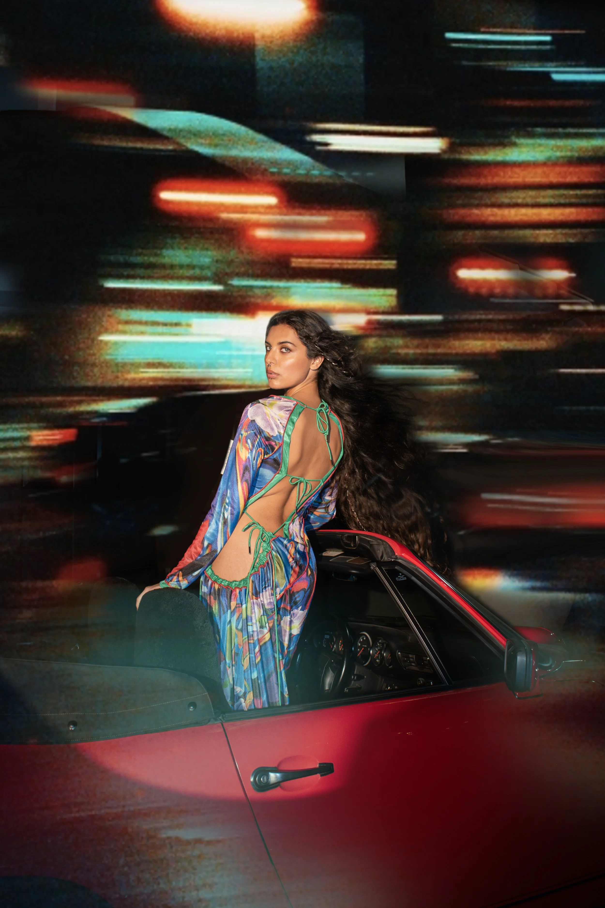 A woman with long dark hair wearing a colorful, cut-out, flowy dress standing next to a red convertible car at night, with streaks of colorful lights in the background.
