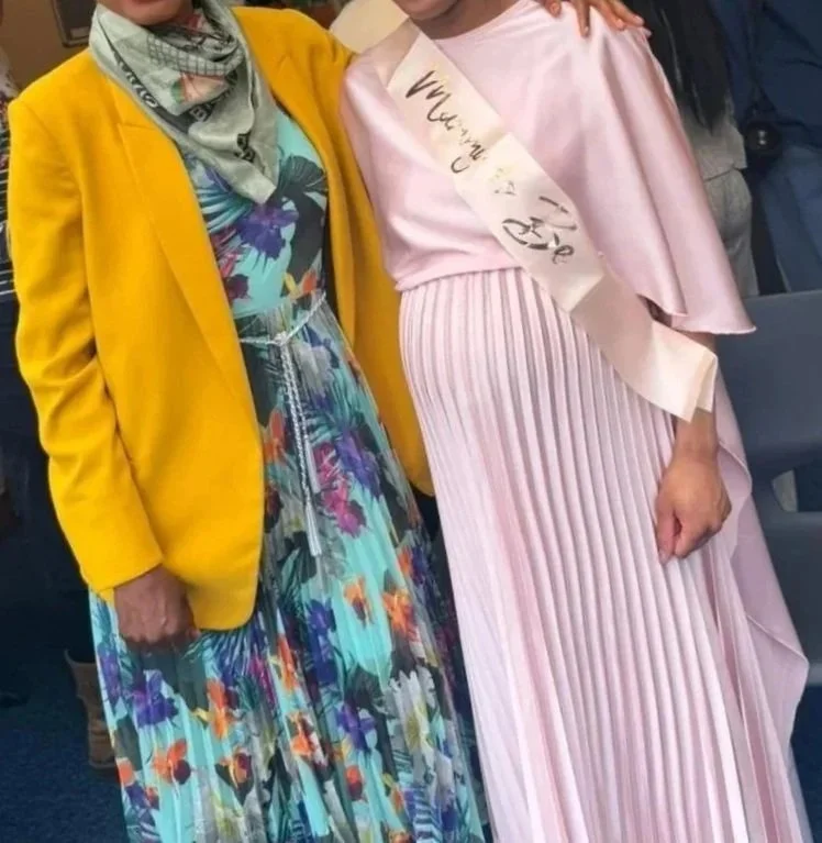 Two women standing close together, one wearing a yellow blazer and a colorful floral dress, the other wearing a pink pleated dress with a sash.