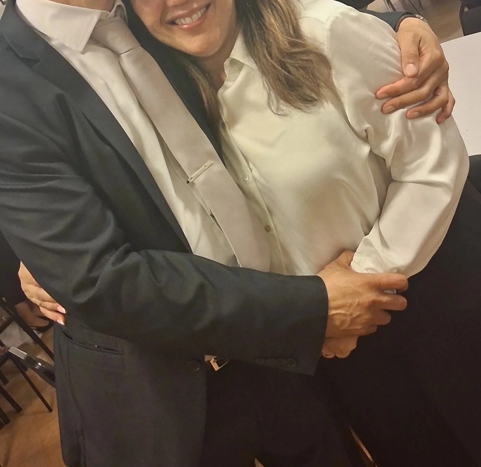 Close-up of a happy woman and a man hugging in a social setting, both smiling, with the woman wearing a white blouse and the man in a dark jacket and light shirt.