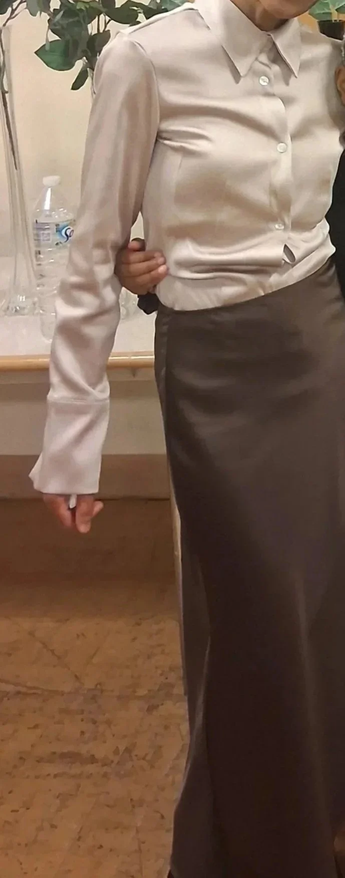 A woman wearing a cream-colored button-up shirt and a dark brown skirt, standing indoors near a table with a water bottle and plant in the background.