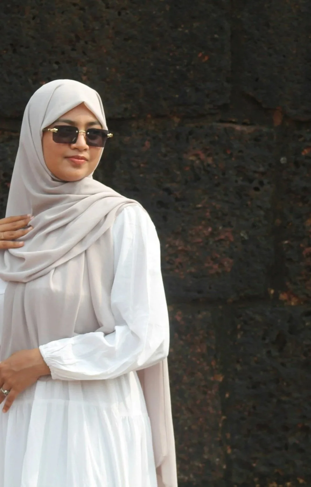 A woman wearing a beige hijab, dark sunglasses, and a white dress, standing in front of a dark, textured background.