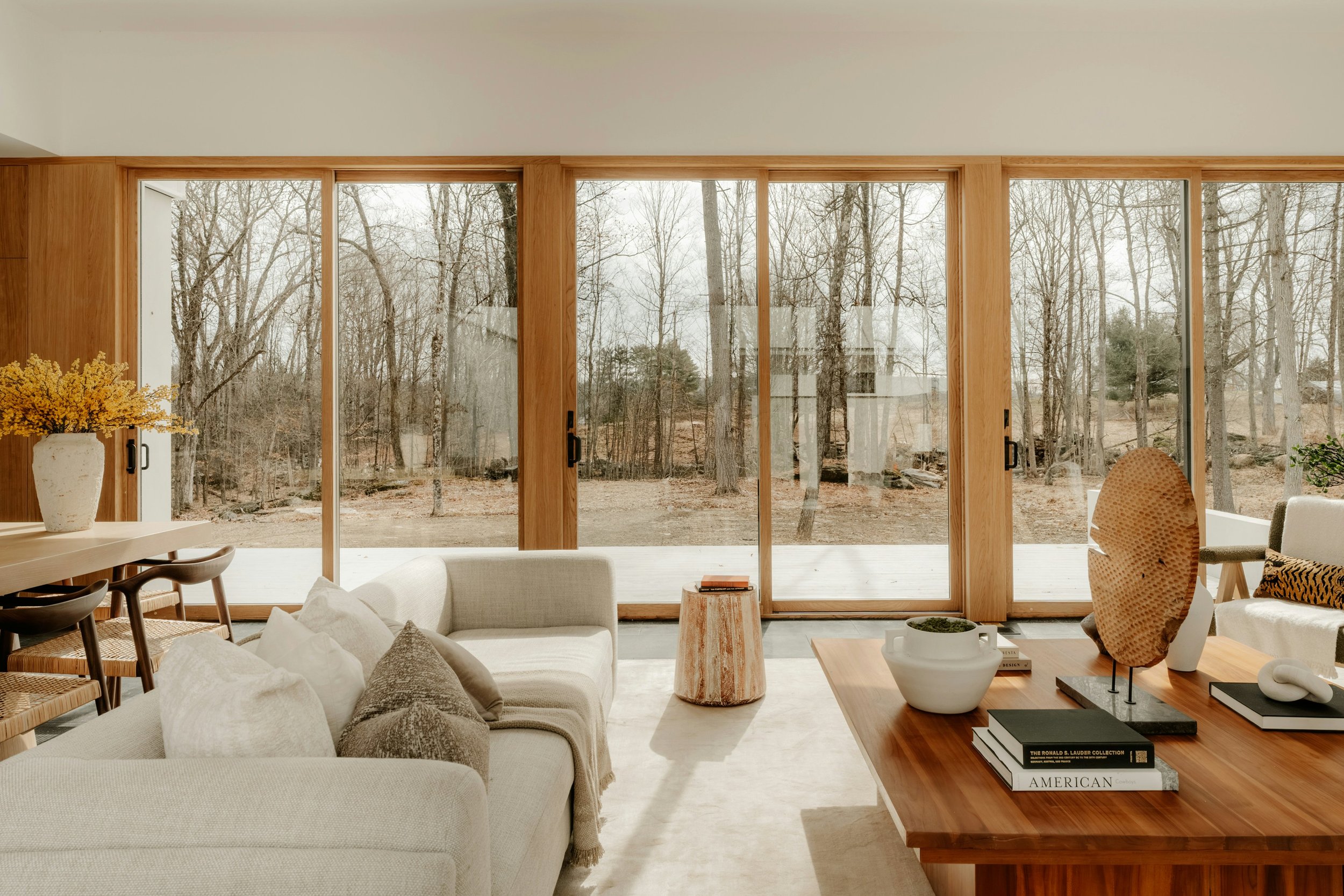 Intérieur moderne d'une maison avec vitres donnant sur un paysage forestier en hiver et mobilier en bois clair et blanc.
