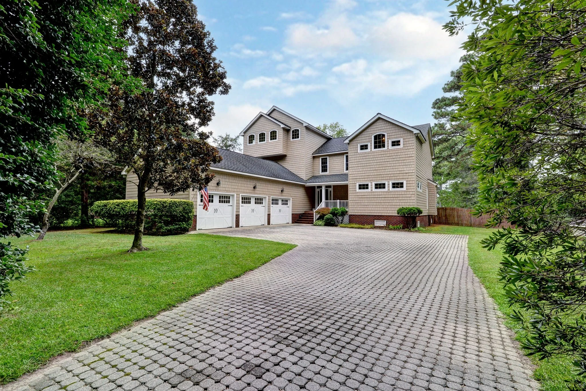 A large house with multiple stories and a three-car garage, set in a green yard with trees and bushes, and a paved driveway.
