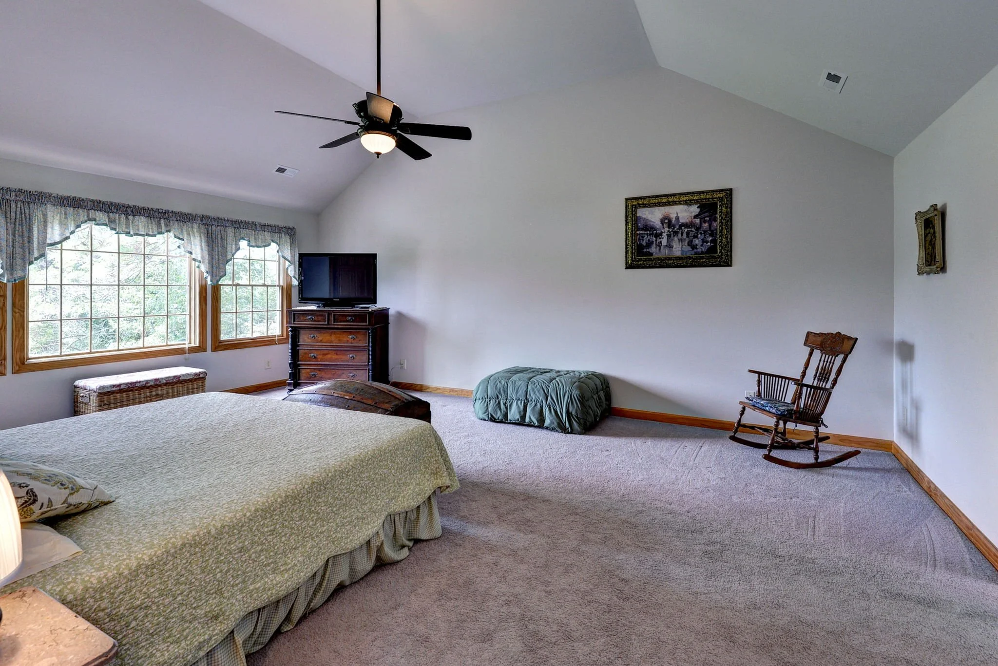 A spacious bedroom with a bed, window seat, dresser, rocking chair, and framed artworks on the wall.