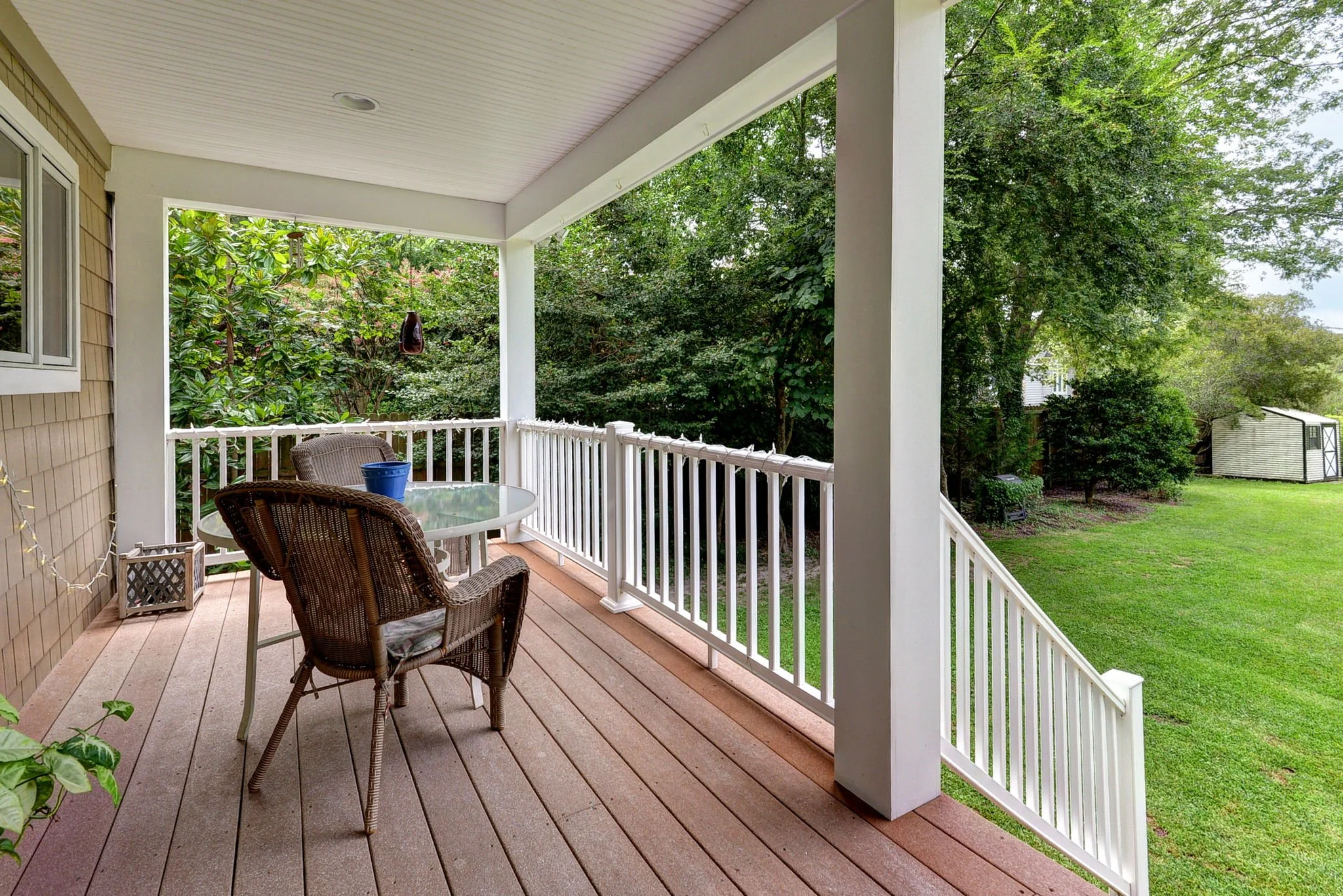 Back porch with outdoor table and wicker chairs overlooking a lush backyard with trees and a shed.