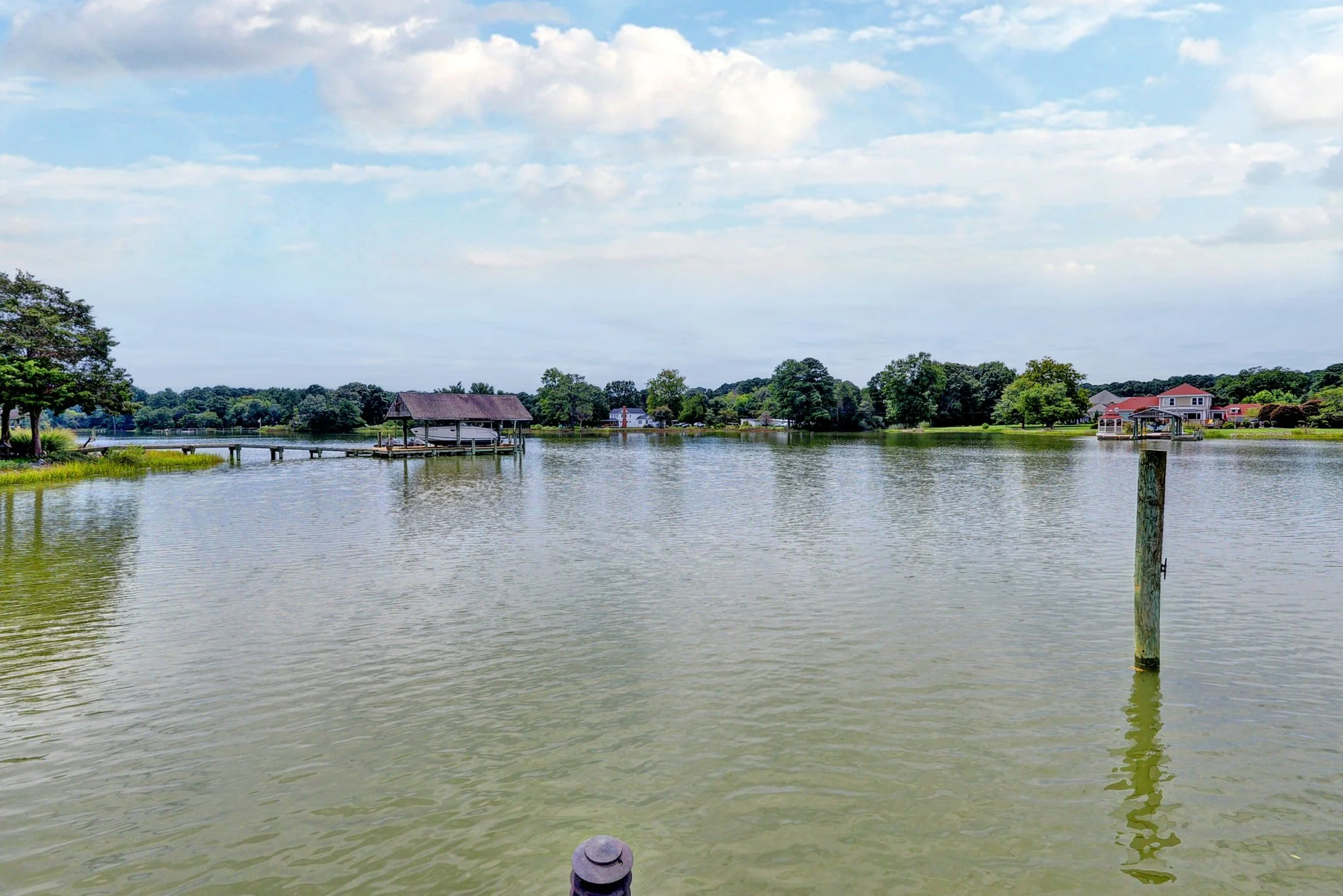 Calm lake with a dock and covered boat shelter on the left, a single wooden post in the water on the right, and trees and houses in the background under a partly cloudy sky.