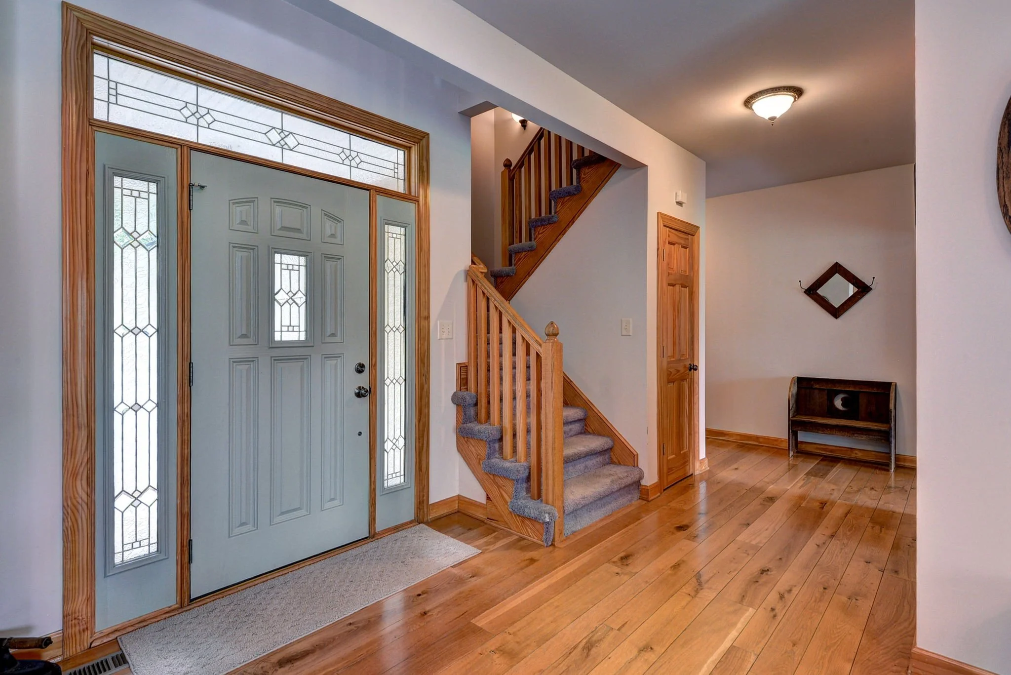 Entryway with front door, glass sidelights, wooden flooring, staircase with carpet, and small console table.
